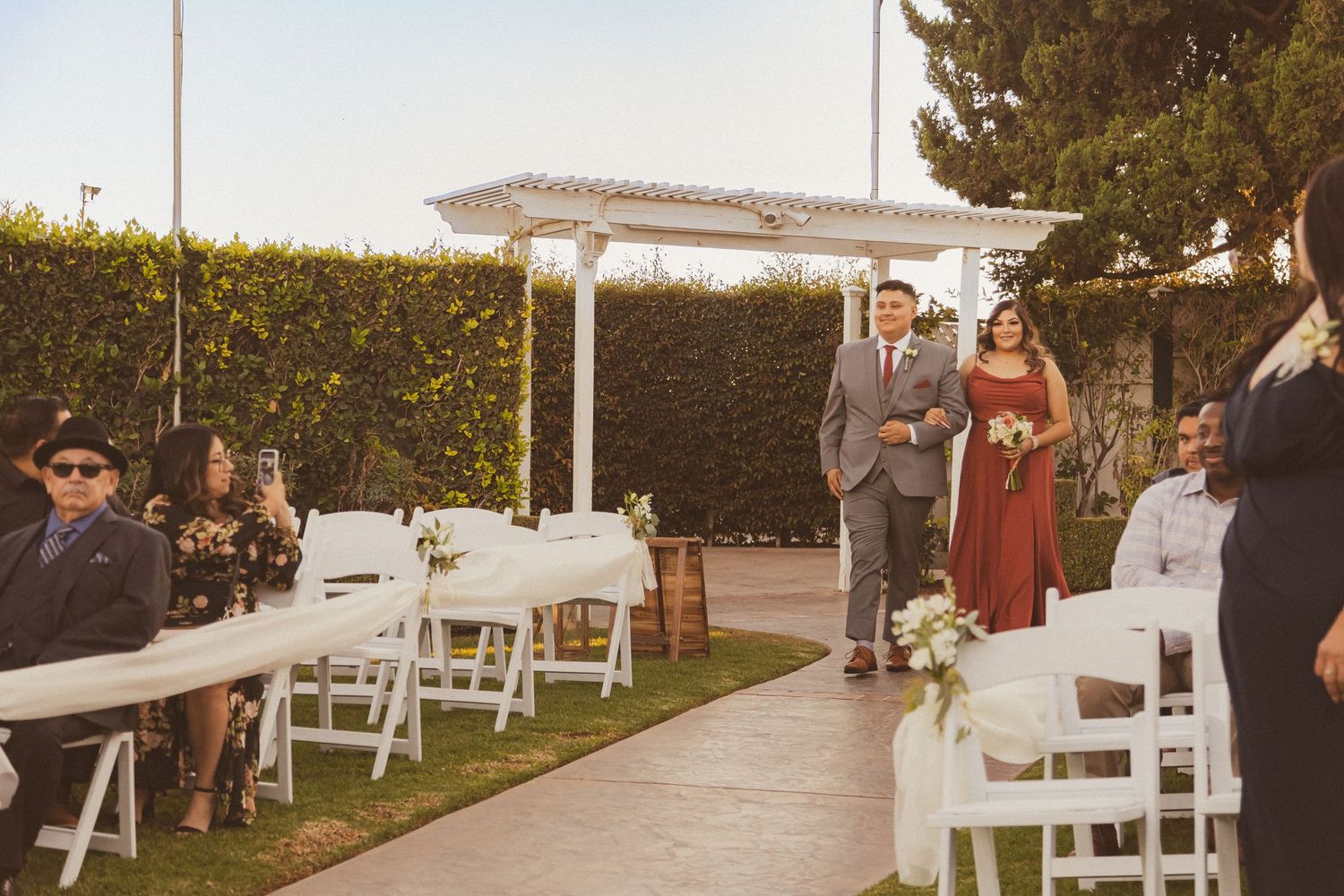 Outdoor garden wedding ceremony setup with white chairs and pergola against a hedge wall at sunset.