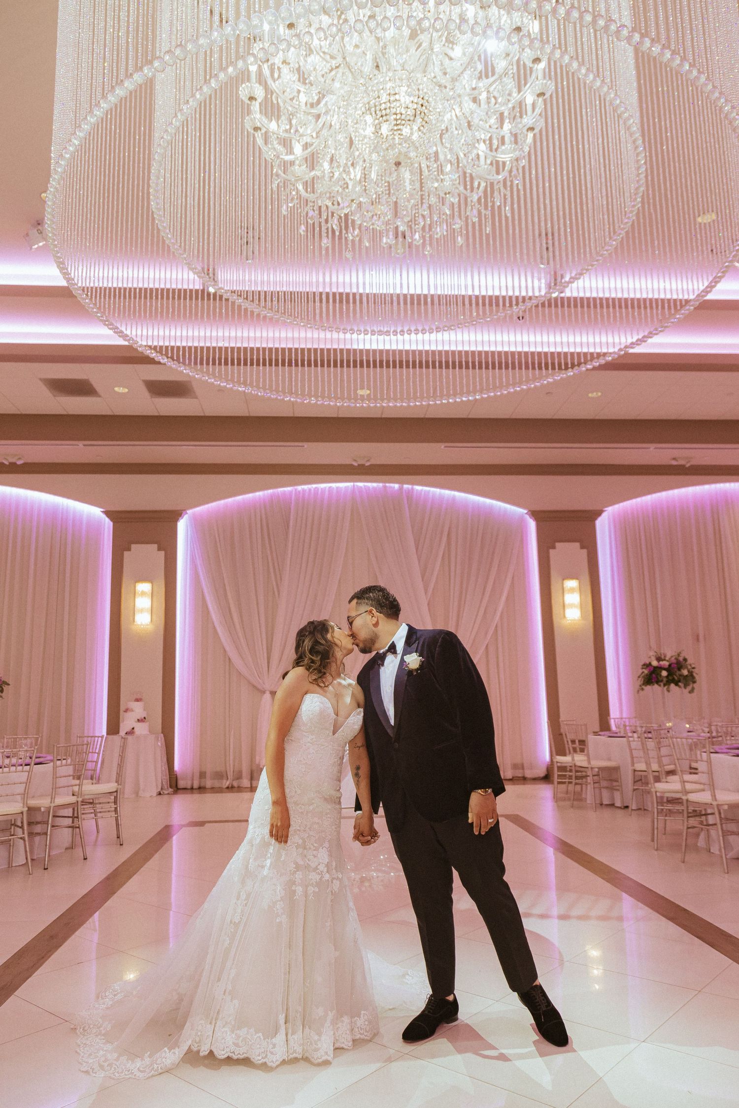 Romantic wedding moment captured in a pink-lit ballroom beneath a stunning crystal chandelier.