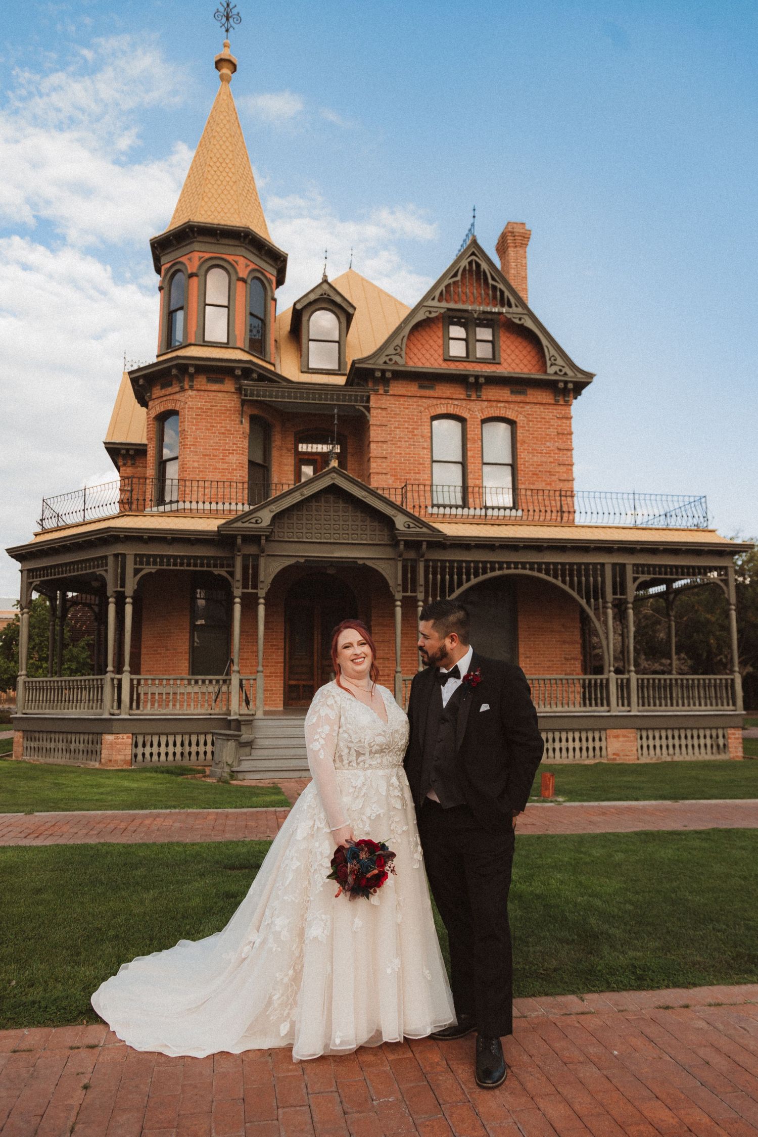 A wedding portrait set against a beautiful red brick Victorian mansion with distinctive architectural features.