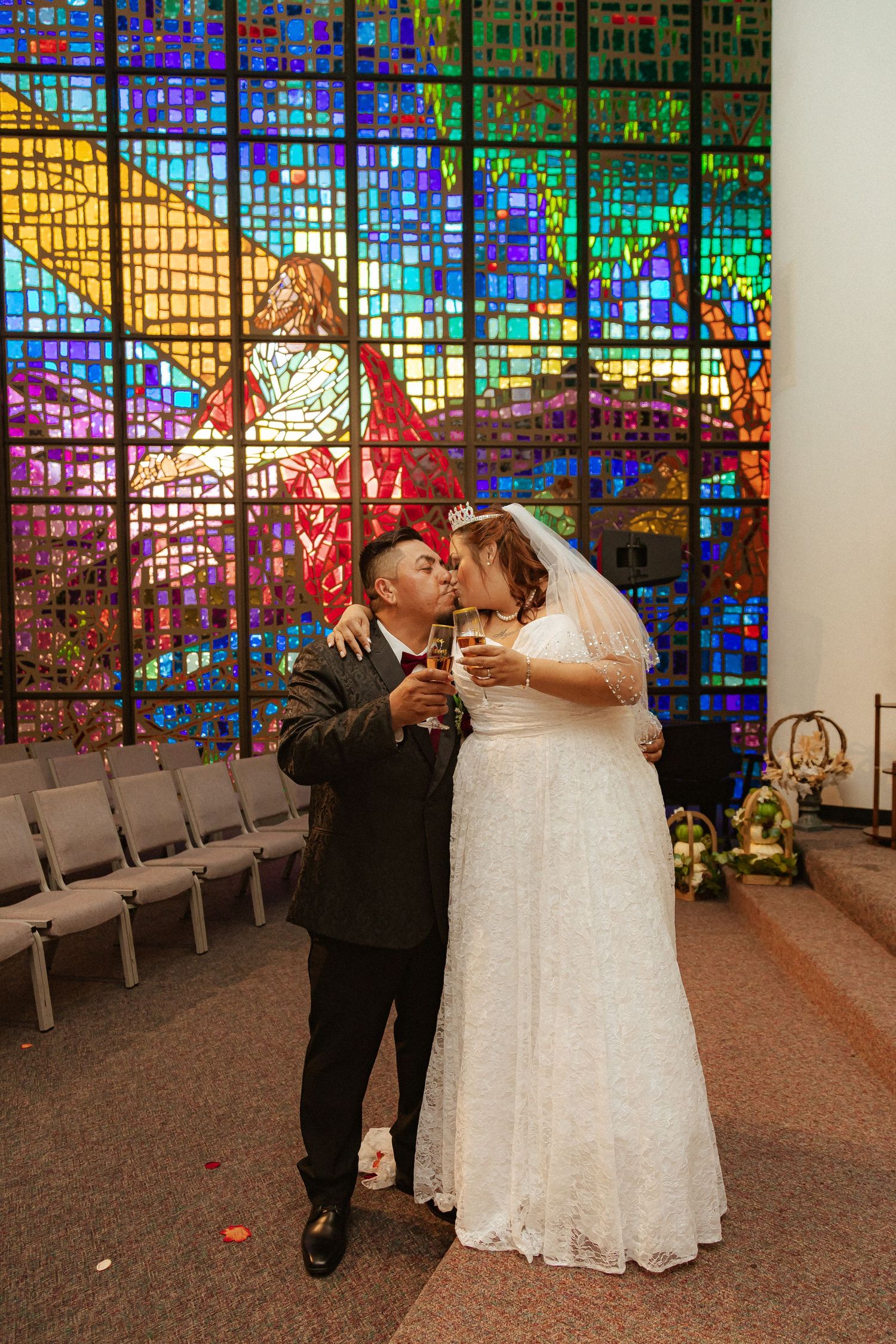 Couple toasts with drinks in front of vibrant stained glass church windows during wedding celebration.