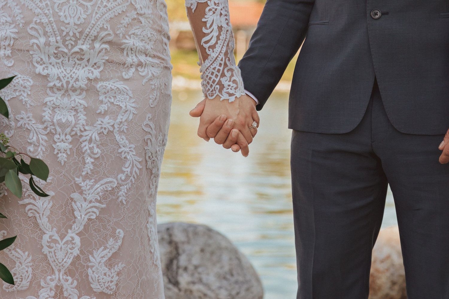 A close-up detail shot of newlyweds holding hands at a waterfront wedding venue.