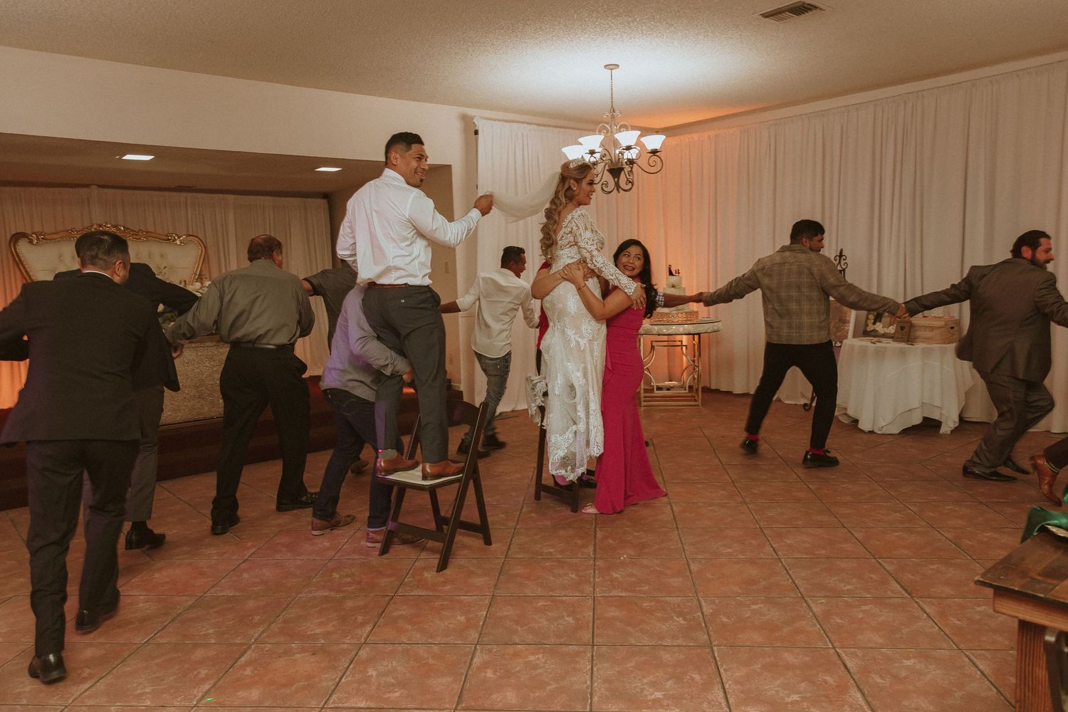 Guests perform a lively group dance routine at a wedding reception with white draped walls.