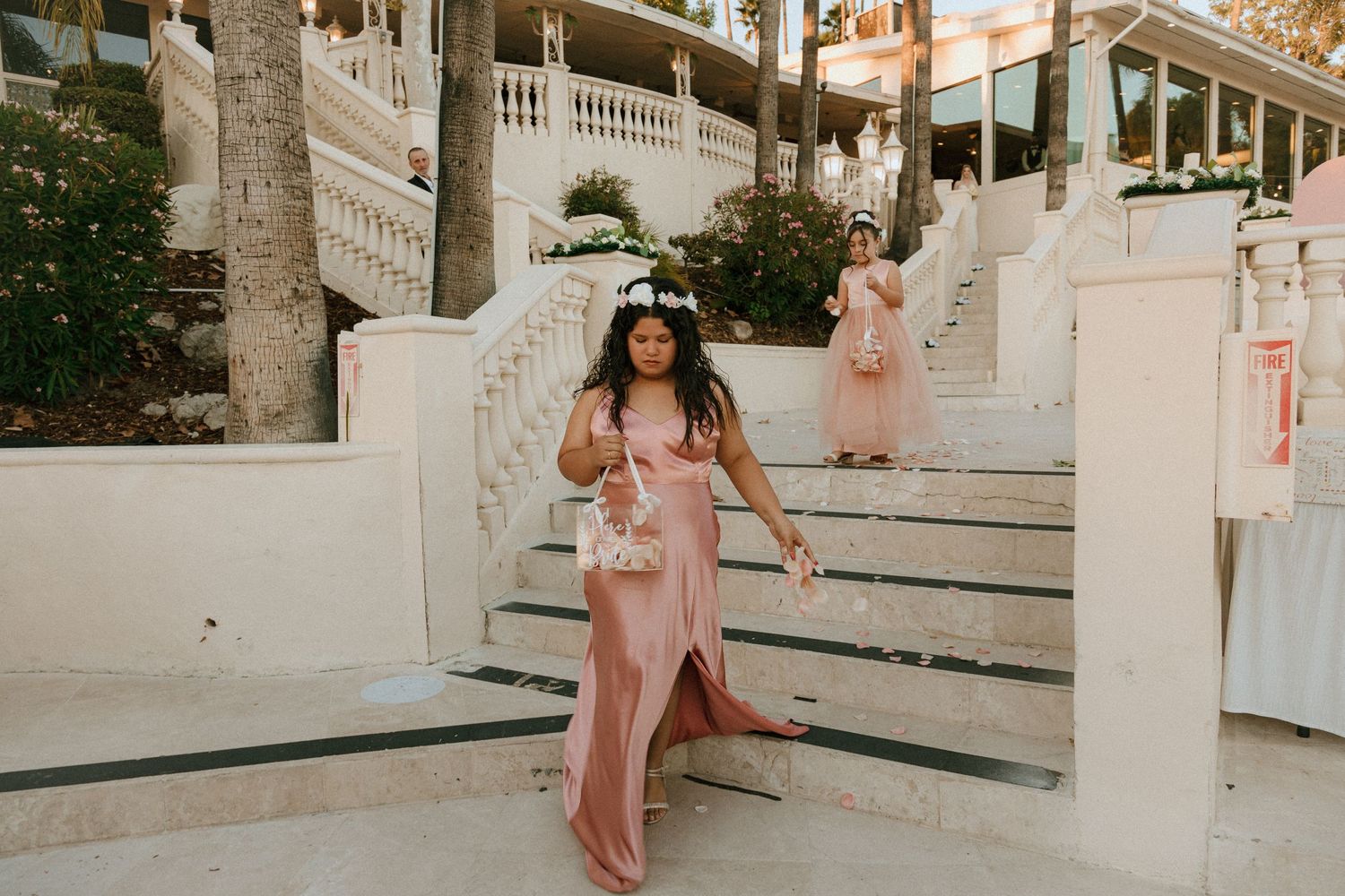 Person in floral crown and pink dress stands on ornate white staircase at sunset.