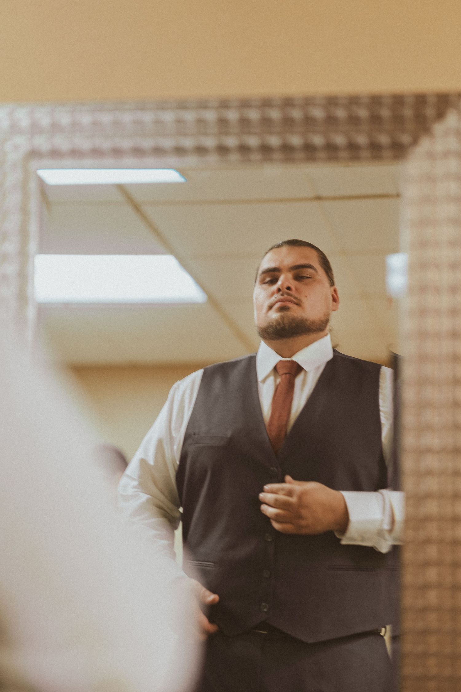 Person wearing dark vest and orange tie looks in mirror while adjusting formal attire.