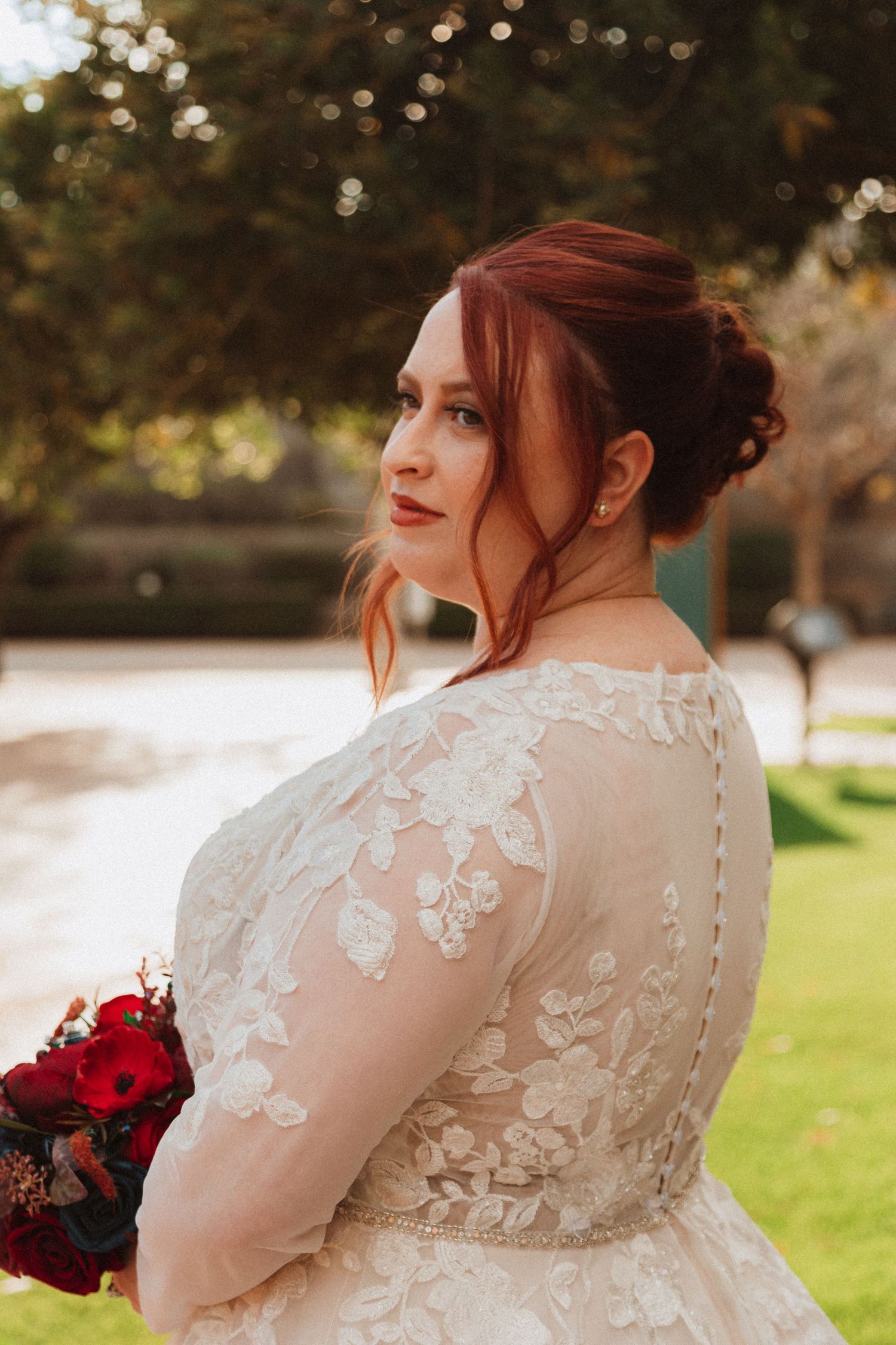 A person in a lace wedding gown with red hair styled up holds a deep red bouquet outdoors.