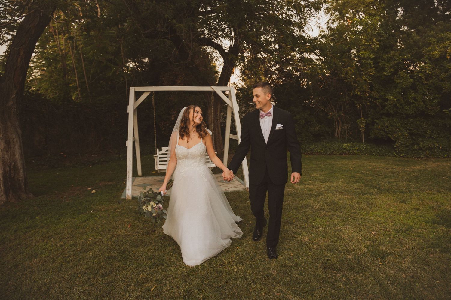 A newly married couple walks together on a grassy lawn with a white wooden arch in the background during golden hour.