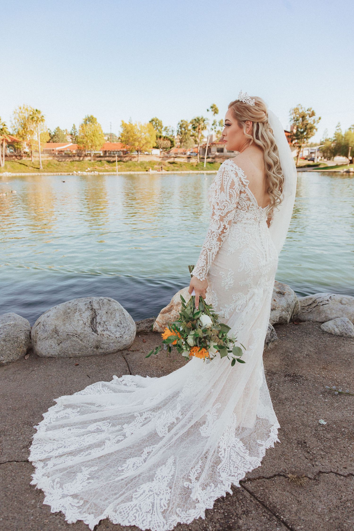 Side profile of lace wedding gown with dramatic train photographed beside lake during golden hour.