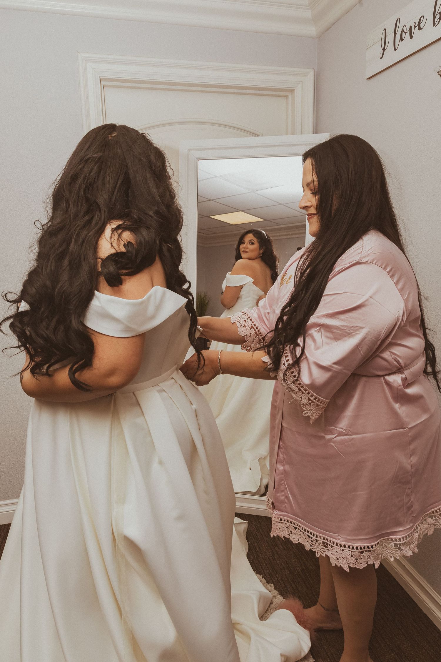 Bridal preparation moment reflected in mirror with pink satin robe and white gown.