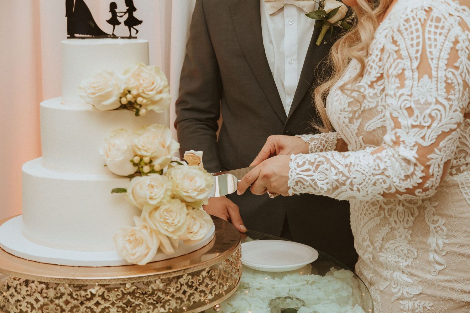 Close-up of hands cutting a white wedding cake decorated with cream roses on an ornate gold stand.