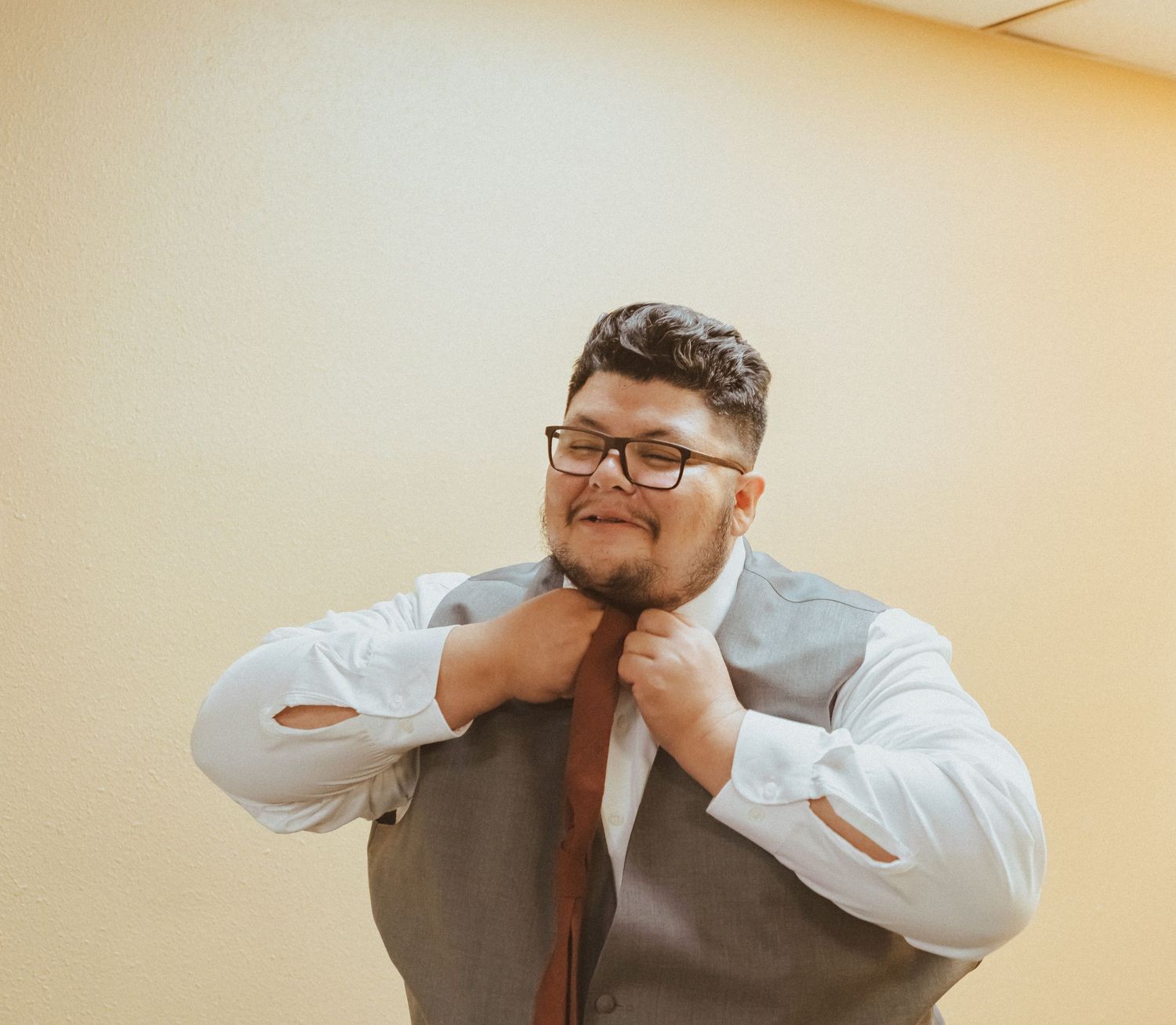A person adjusts their burgundy tie while wearing a white dress shirt and glasses in an indoor setting.