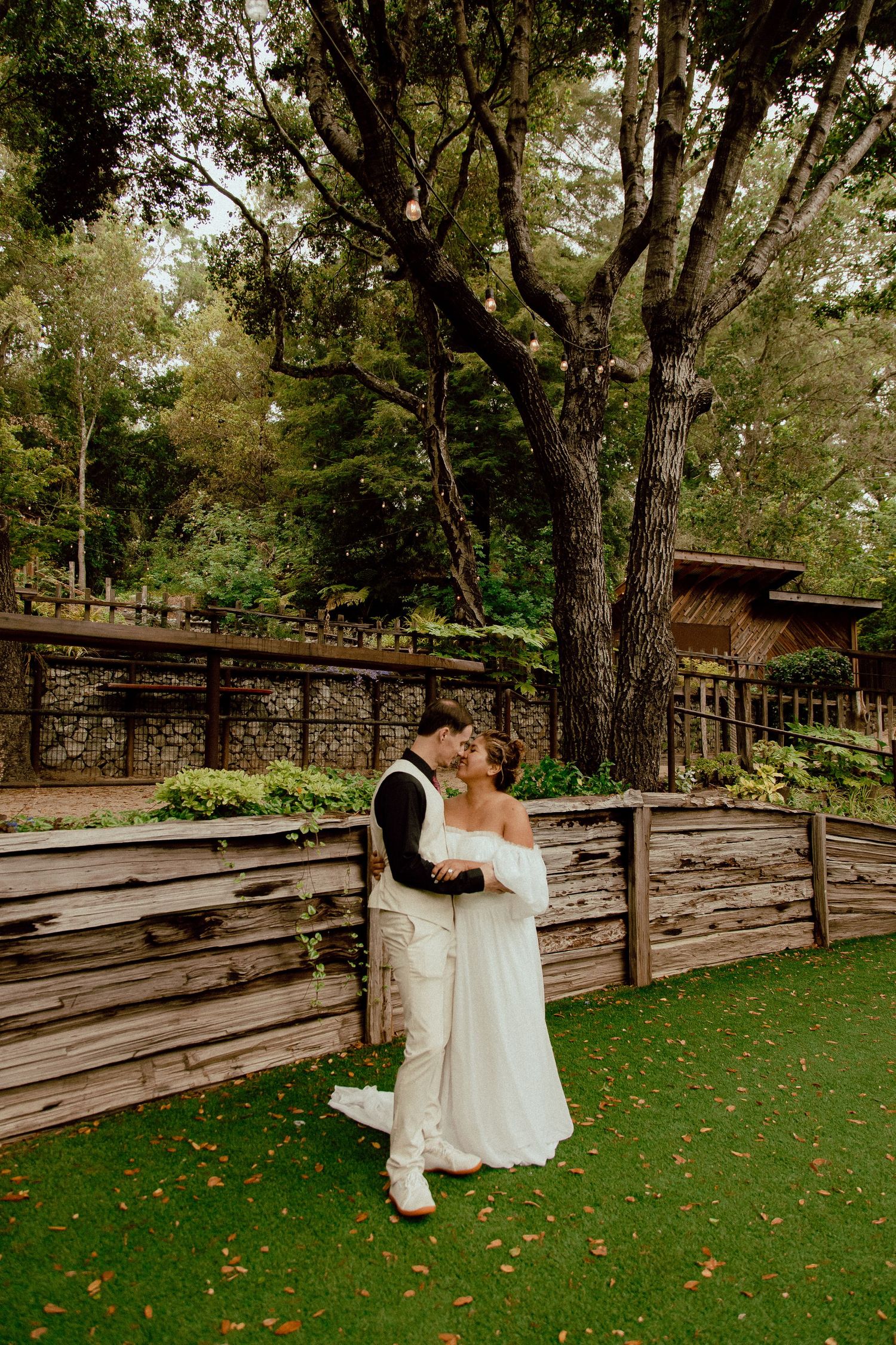 A wedding couple embraces on a lawn in front of a rustic wooden fence and lush trees.