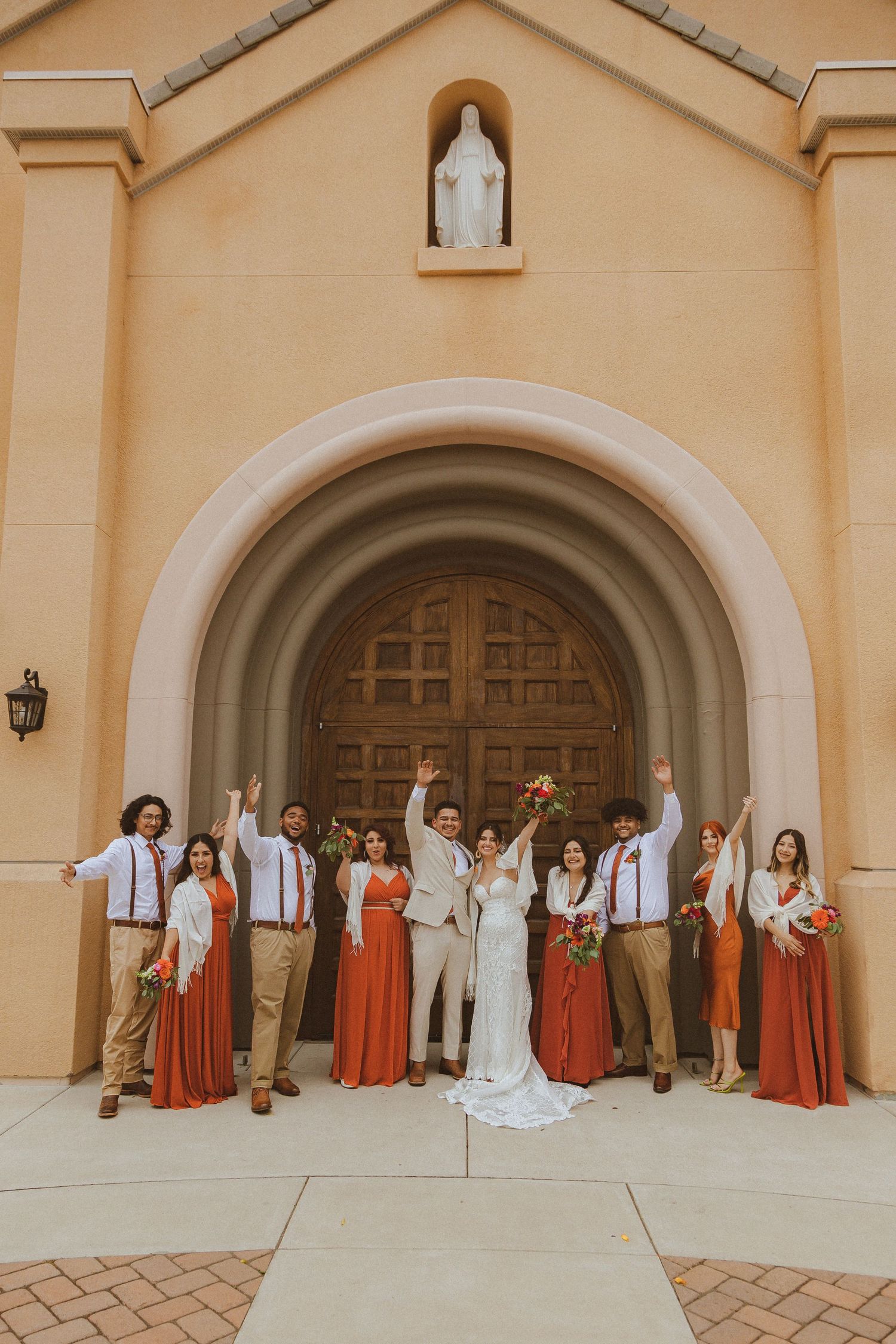 Full wedding party poses enthusiastically in front of church entrance with statue above.