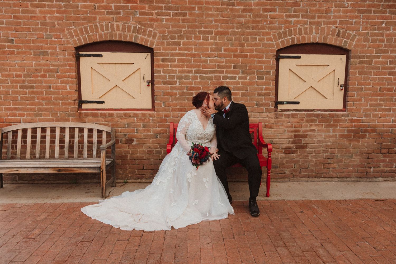 A wedding couple sits intimately on a wooden bench against a rustic brick wall with white wooden shutters.