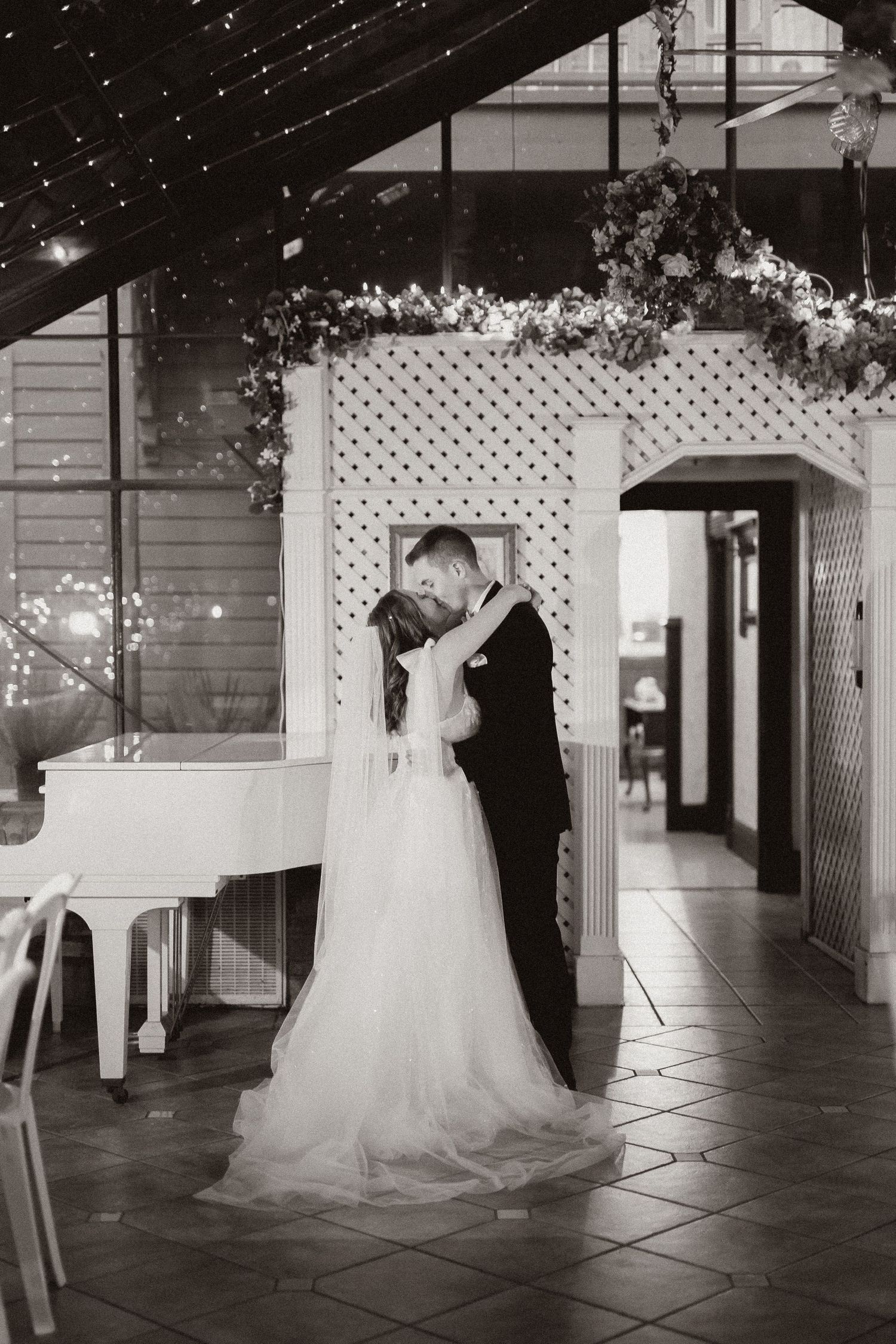 Black and white photo of newlyweds sharing intimate moment under decorated lattice archway beside white piano.