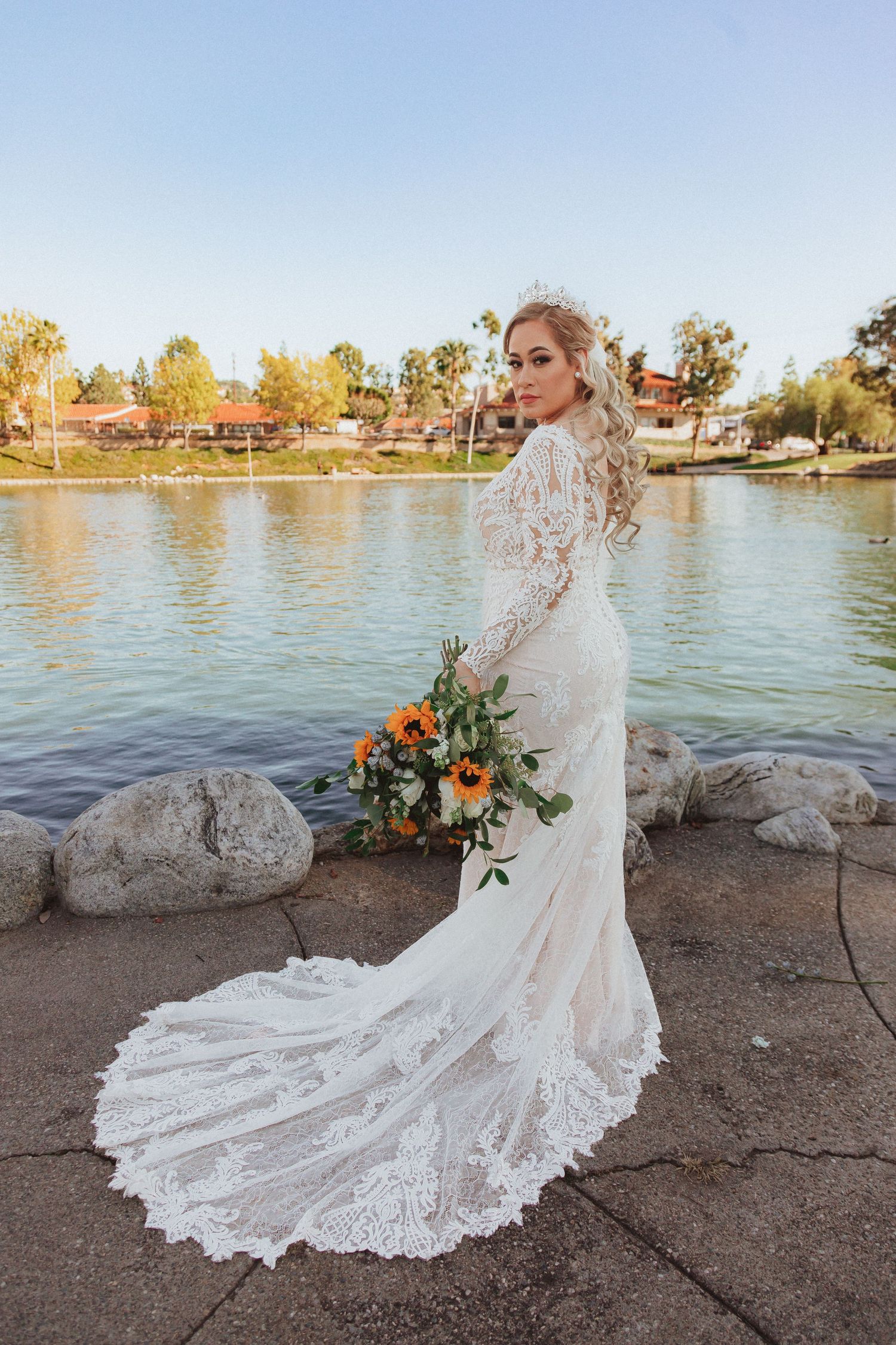 Wedding photography featuring long-sleeved lace gown and cascading bouquet by waterfront.