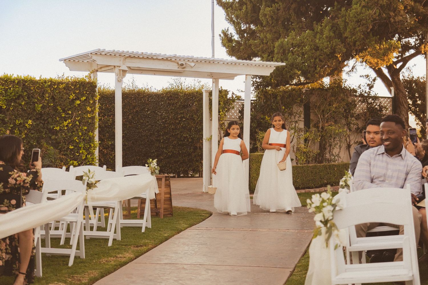Wedding ceremony space with white pergola and flower arrangements along garden path.