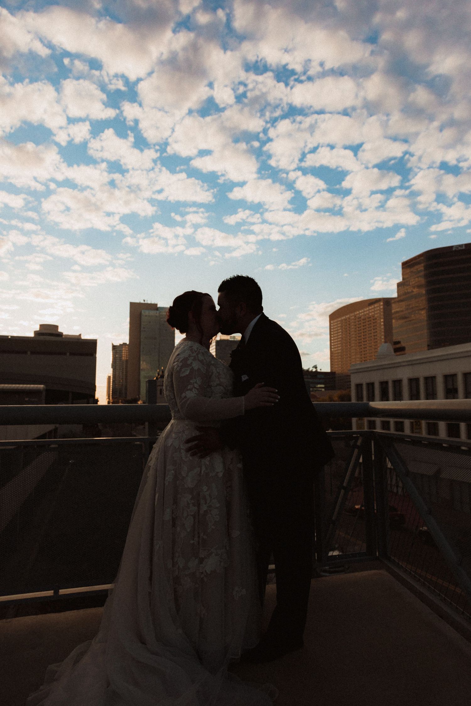 Silhouetted wedding couple against dramatic cloudy sky during sunset on city rooftop.