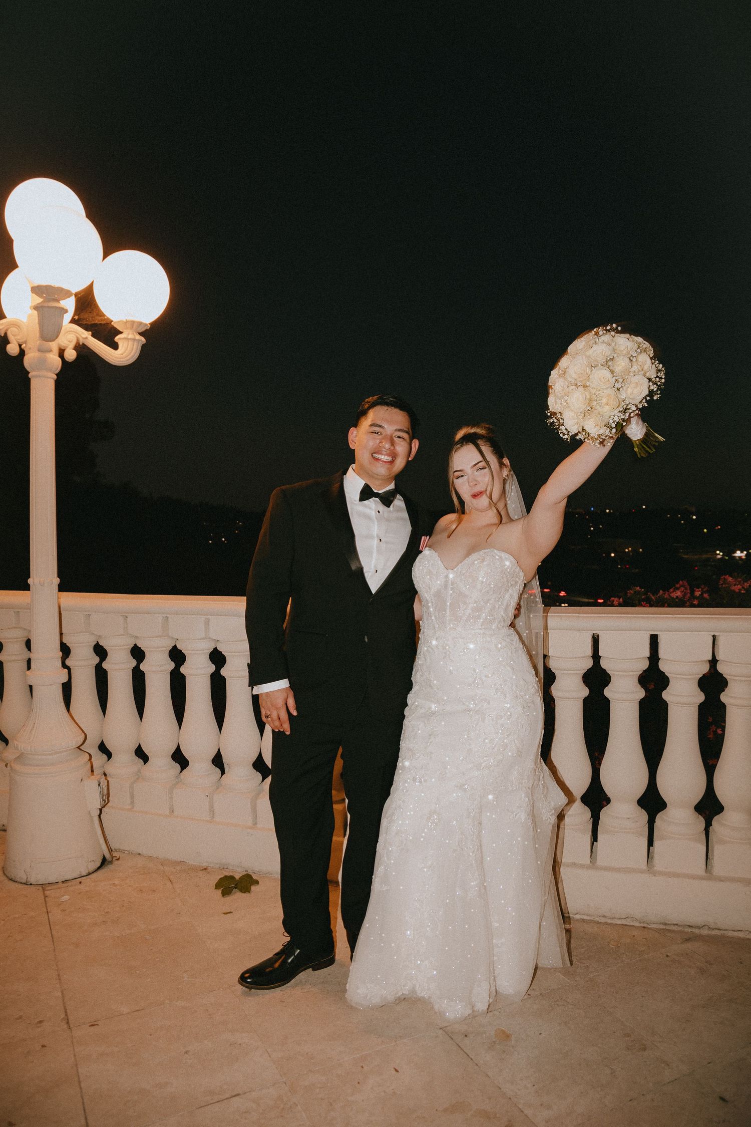 A romantic wedding couple poses on a balcony at night with decorative lighting and white floral bouquet.