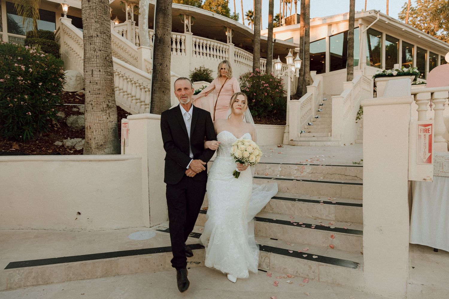 Newlyweds walking down decorative white stairs at an outdoor venue with palm trees in warm lighting.