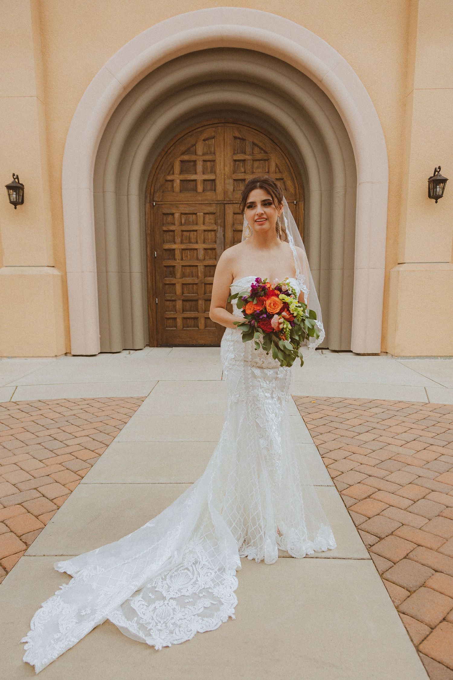 Bride in lace wedding dress holding vibrant orange and red bouquet in front of arched doorway.