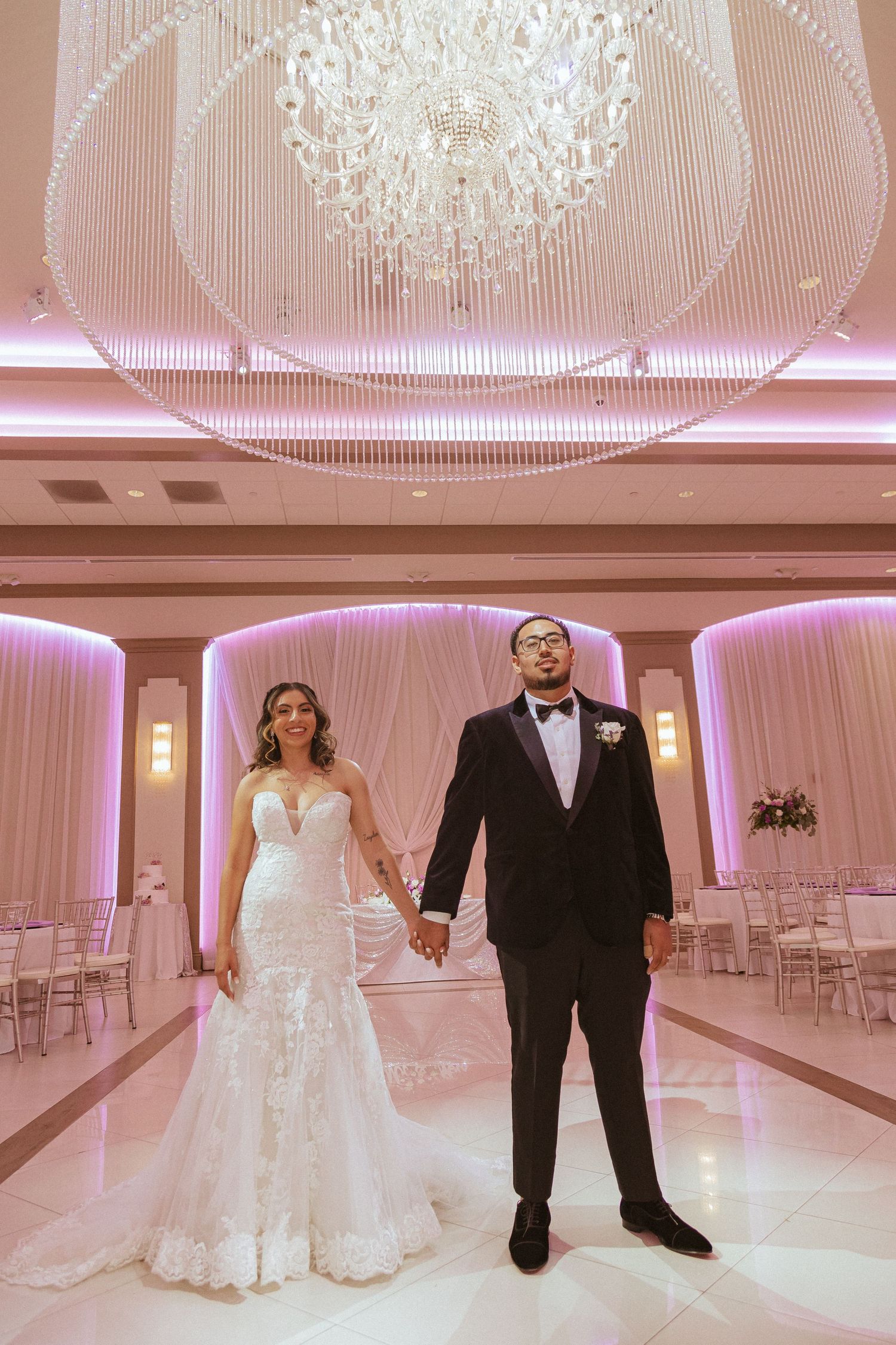Couple holds hands in formal wedding attire under crystal chandelier in pink-lit reception venue.