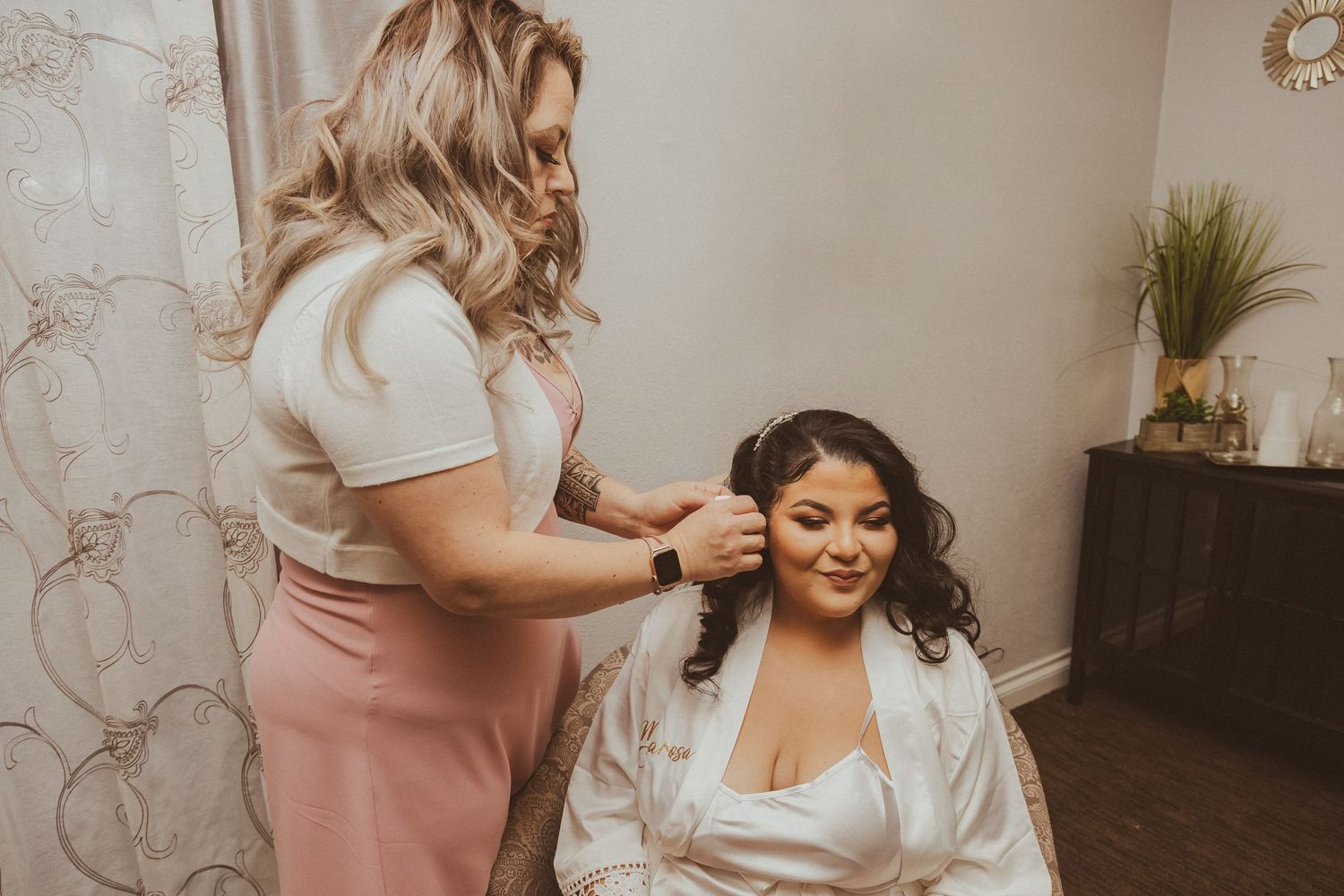 A stylist works on wedding day hair preparations in a room with sheer curtains.