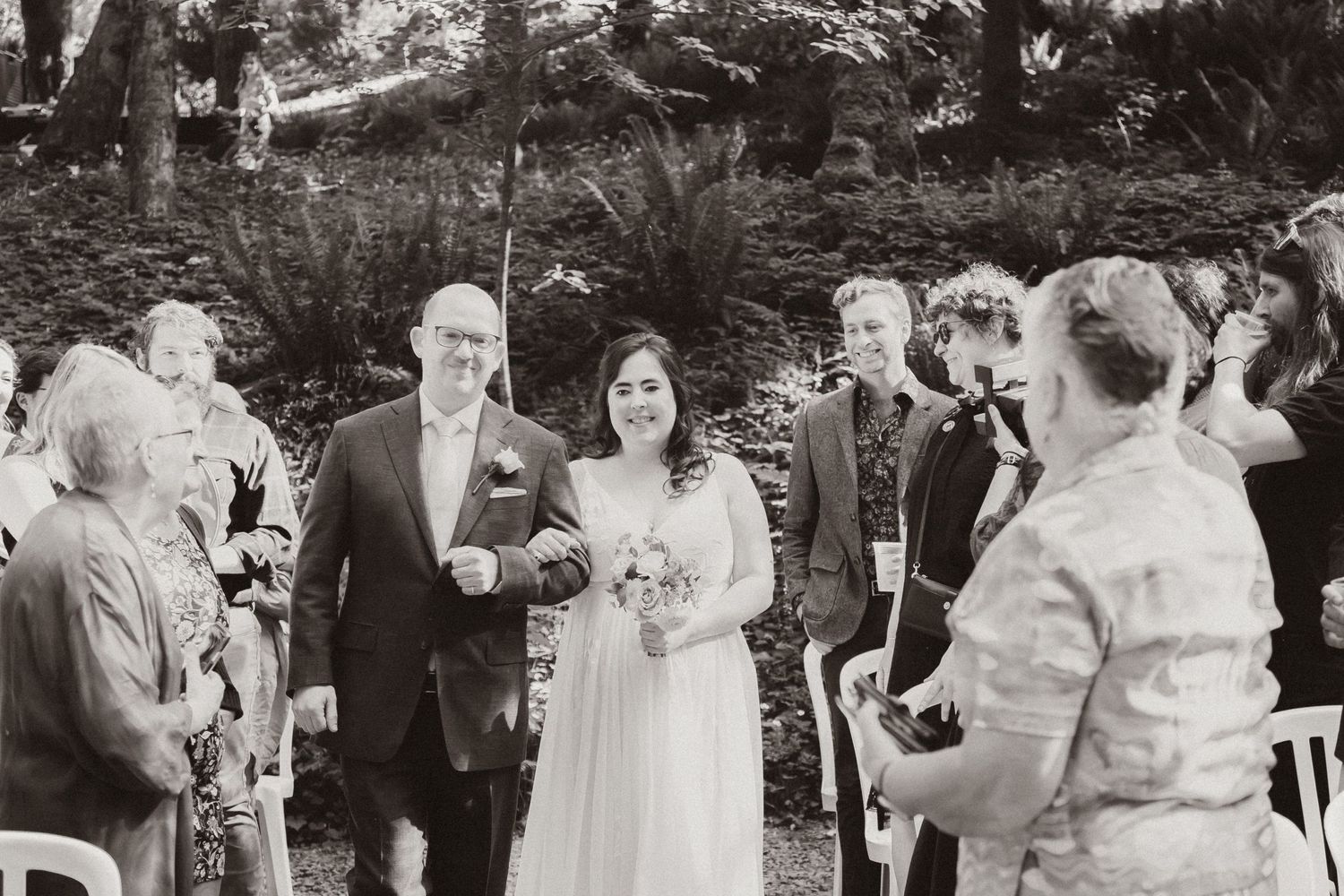 A wedding ceremony processional in a forest setting with guests looking on.