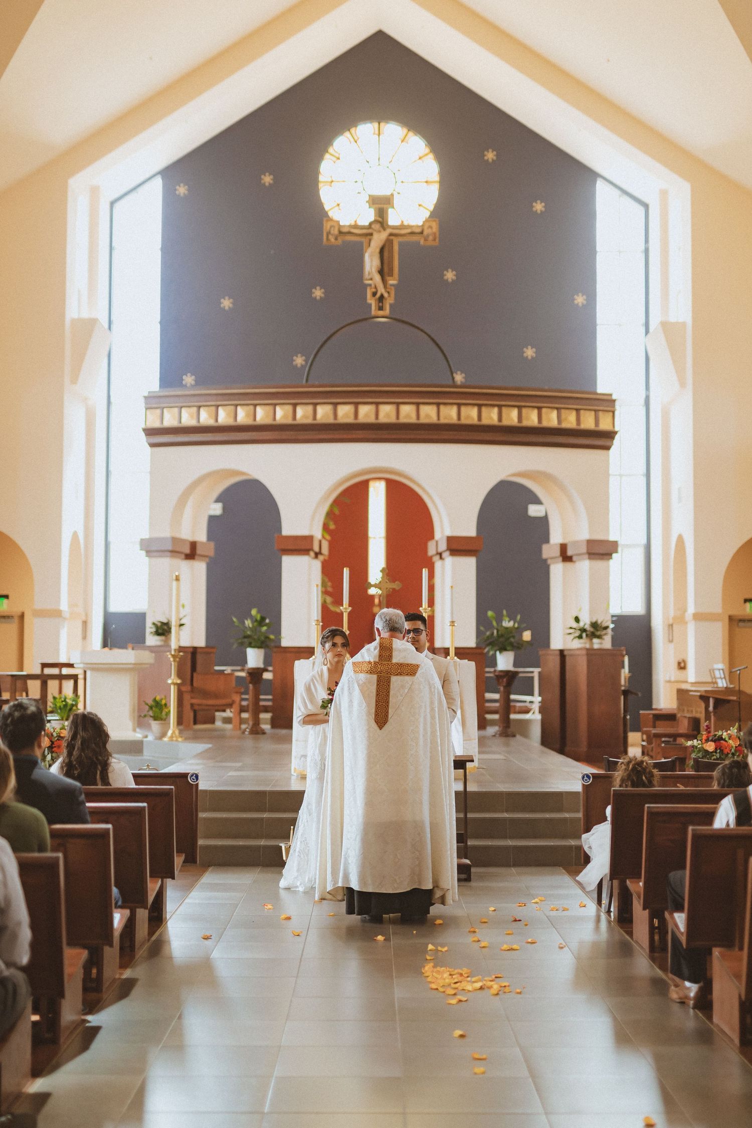 A priest stands at the altar beneath a crucifix during a church ceremony with scattered flower petals.
