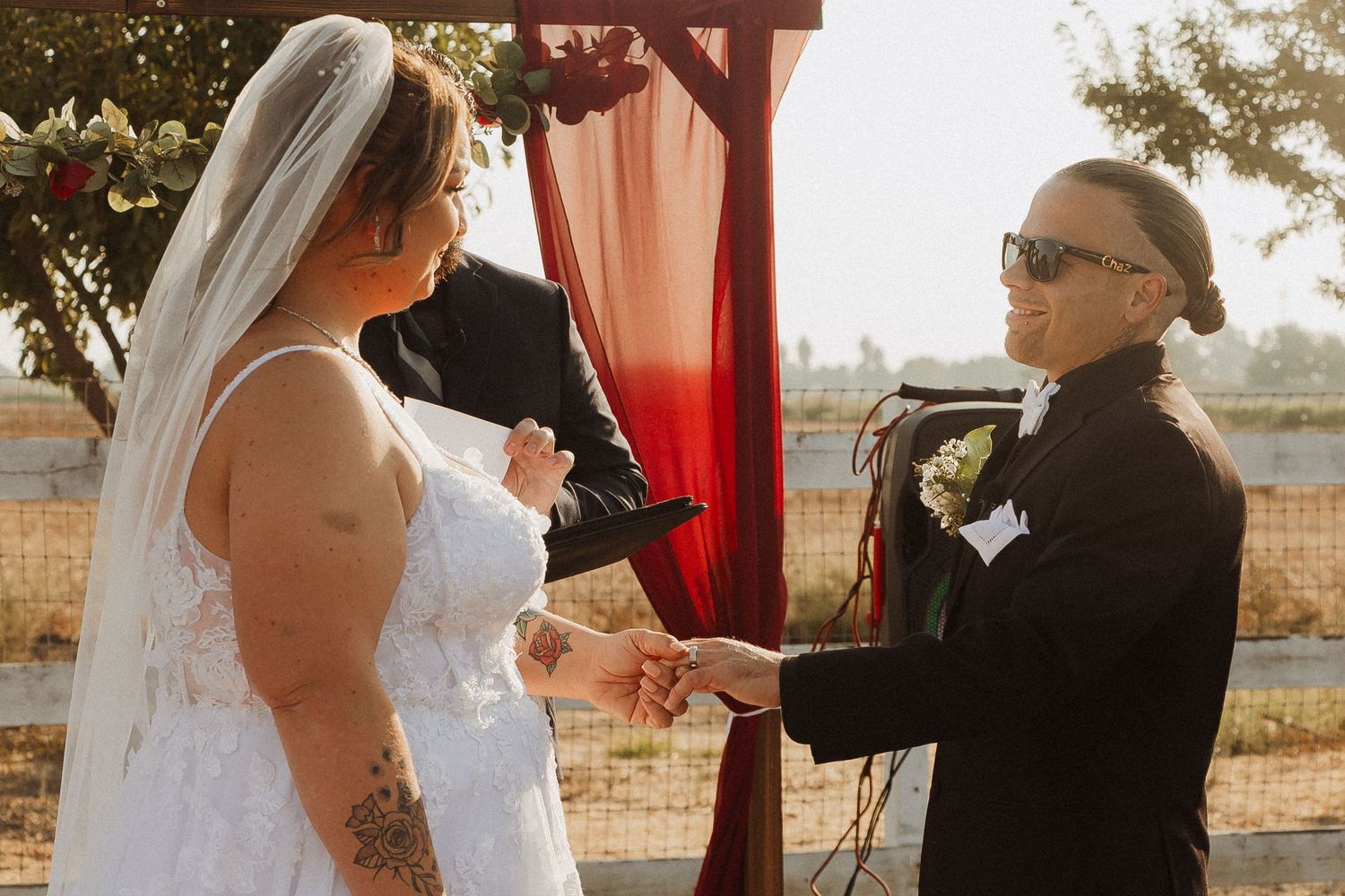 A tender moment during an outdoor wedding ceremony beneath a red-draped archway.