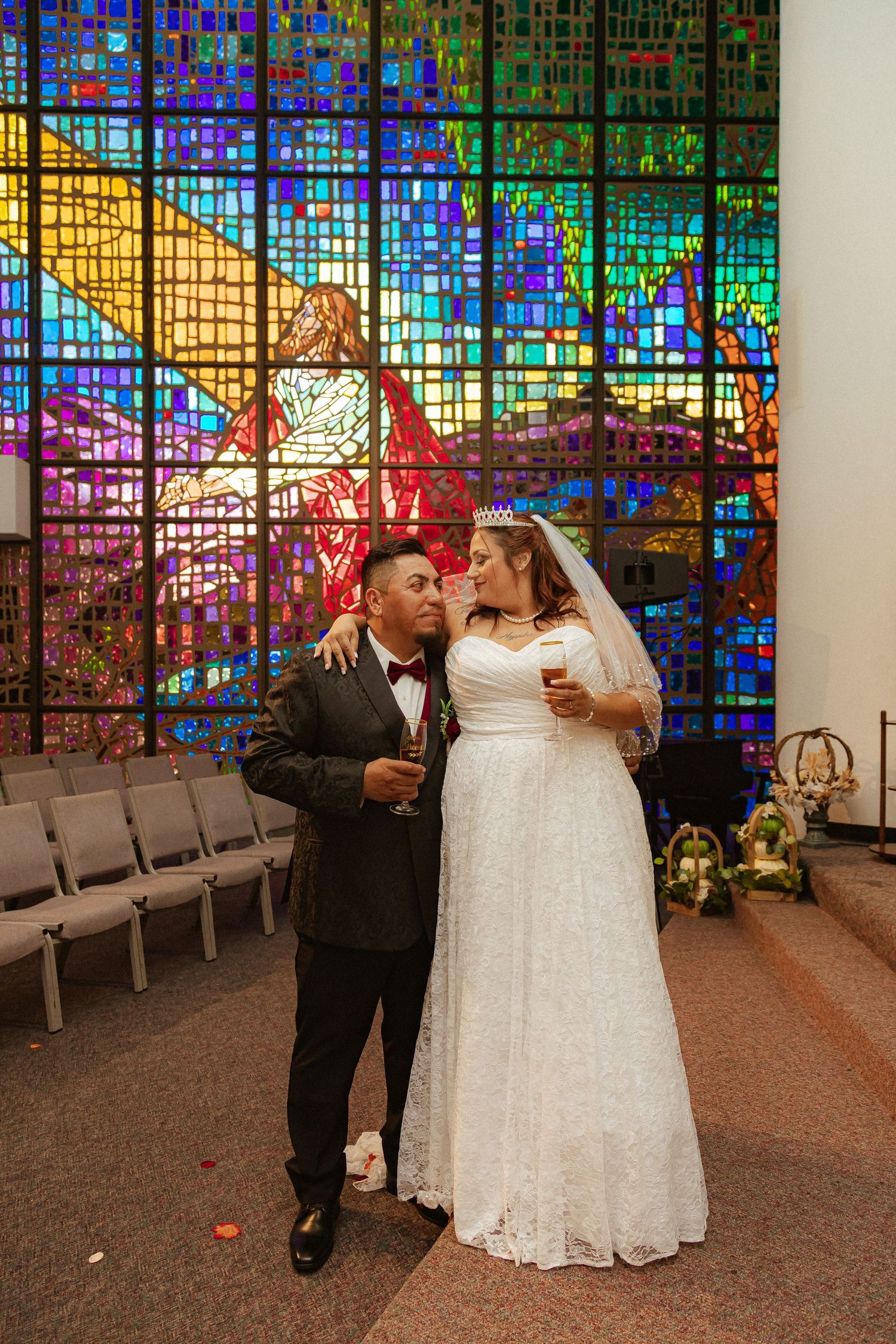 A couple celebrates with champagne in front of vibrant stained glass windows in a church.