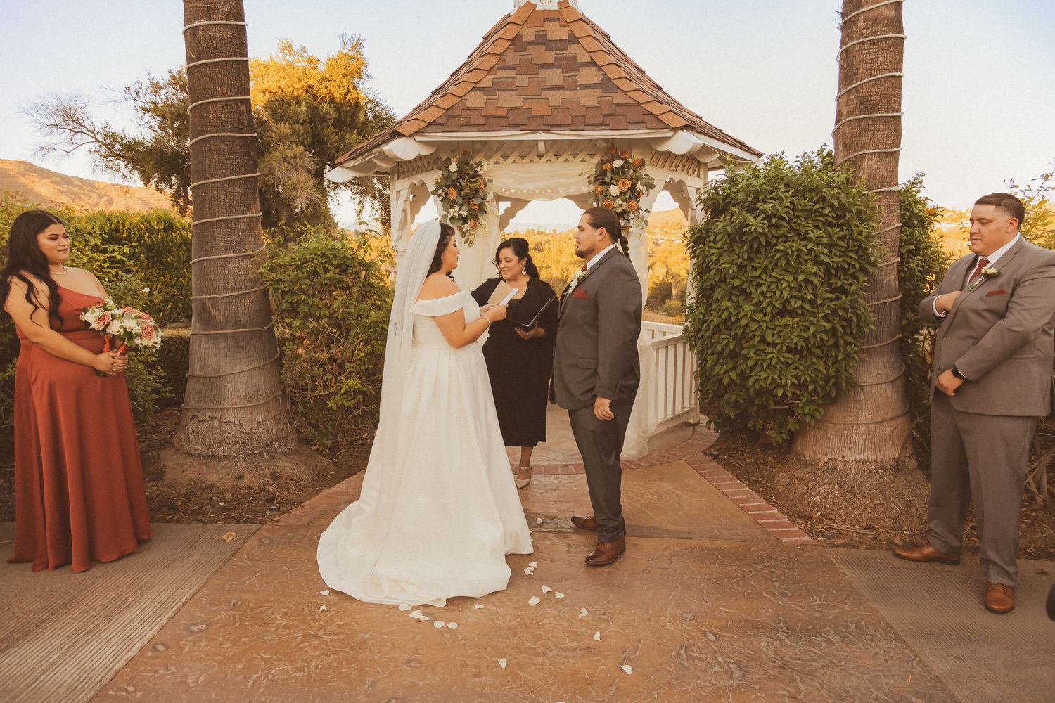 Wedding ceremony in progress under decorated gazebo with autumn colors in background.