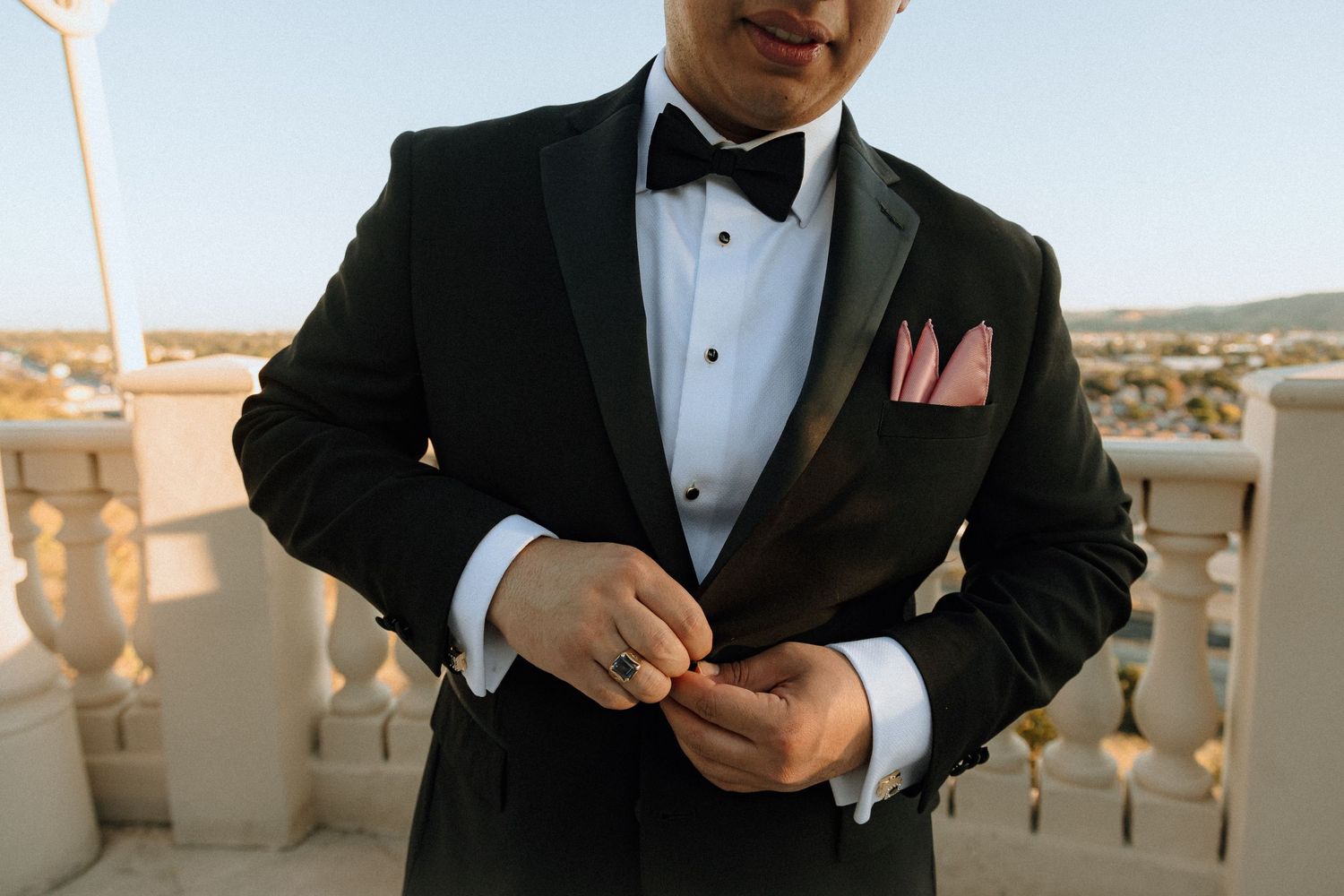 Close-up of groom adjusting cufflinks while wearing classic black tuxedo with pink pocket square.