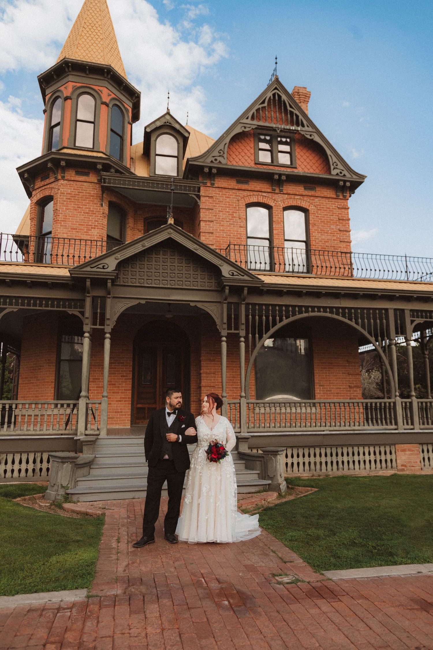 A couple poses in front of a historic red brick Victorian mansion with detailed woodwork and wraparound porch.
