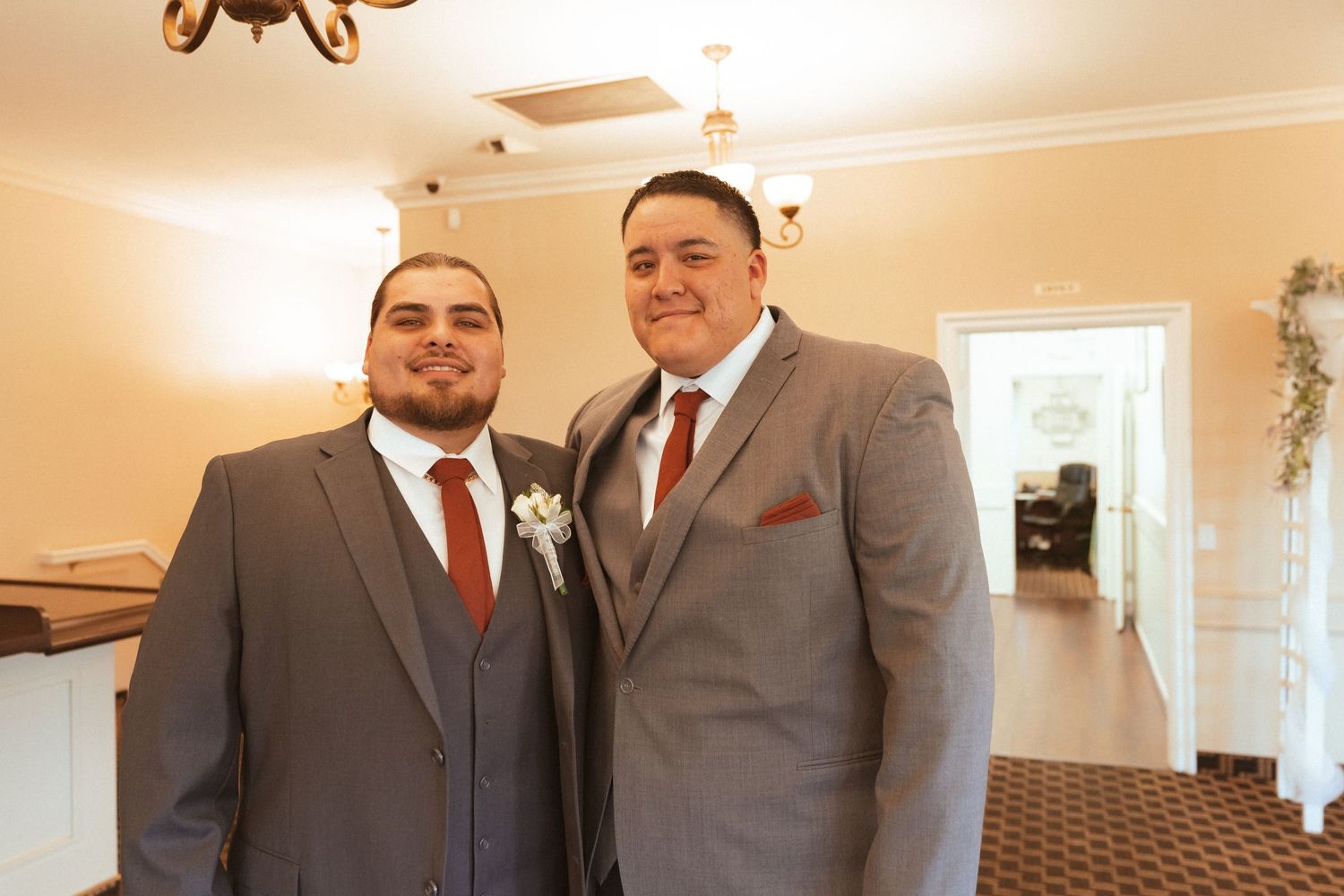 Two groomsmen in matching gray suits and burgundy ties pose together at a wedding venue.