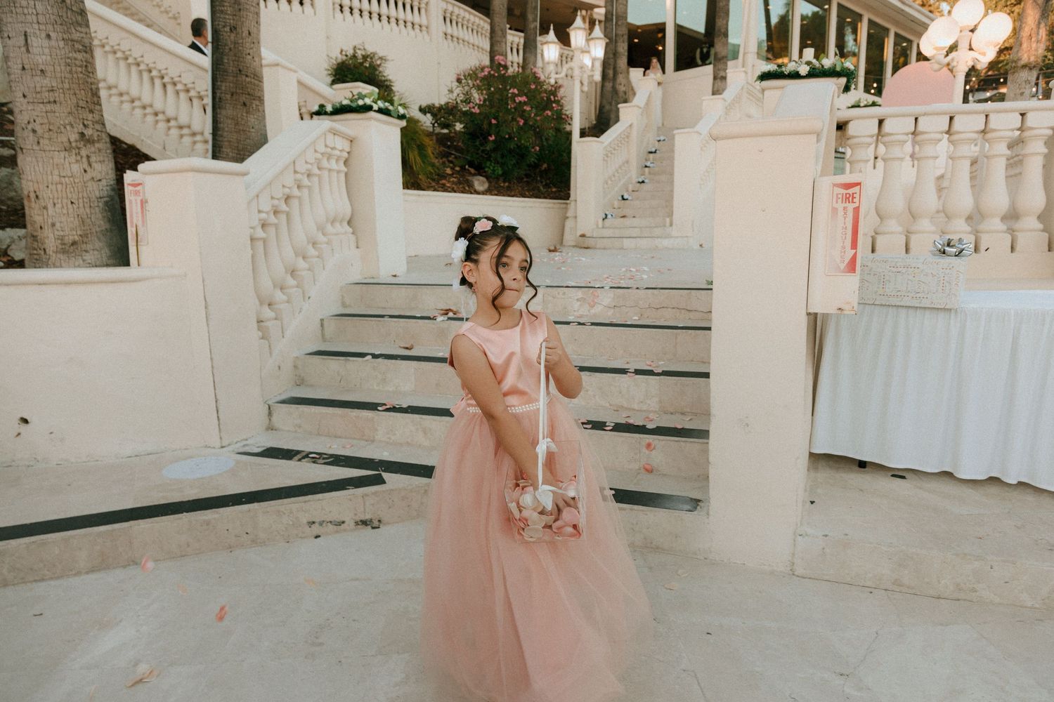 Person in delicate pink gown stands elegantly on white stone steps at dusk.