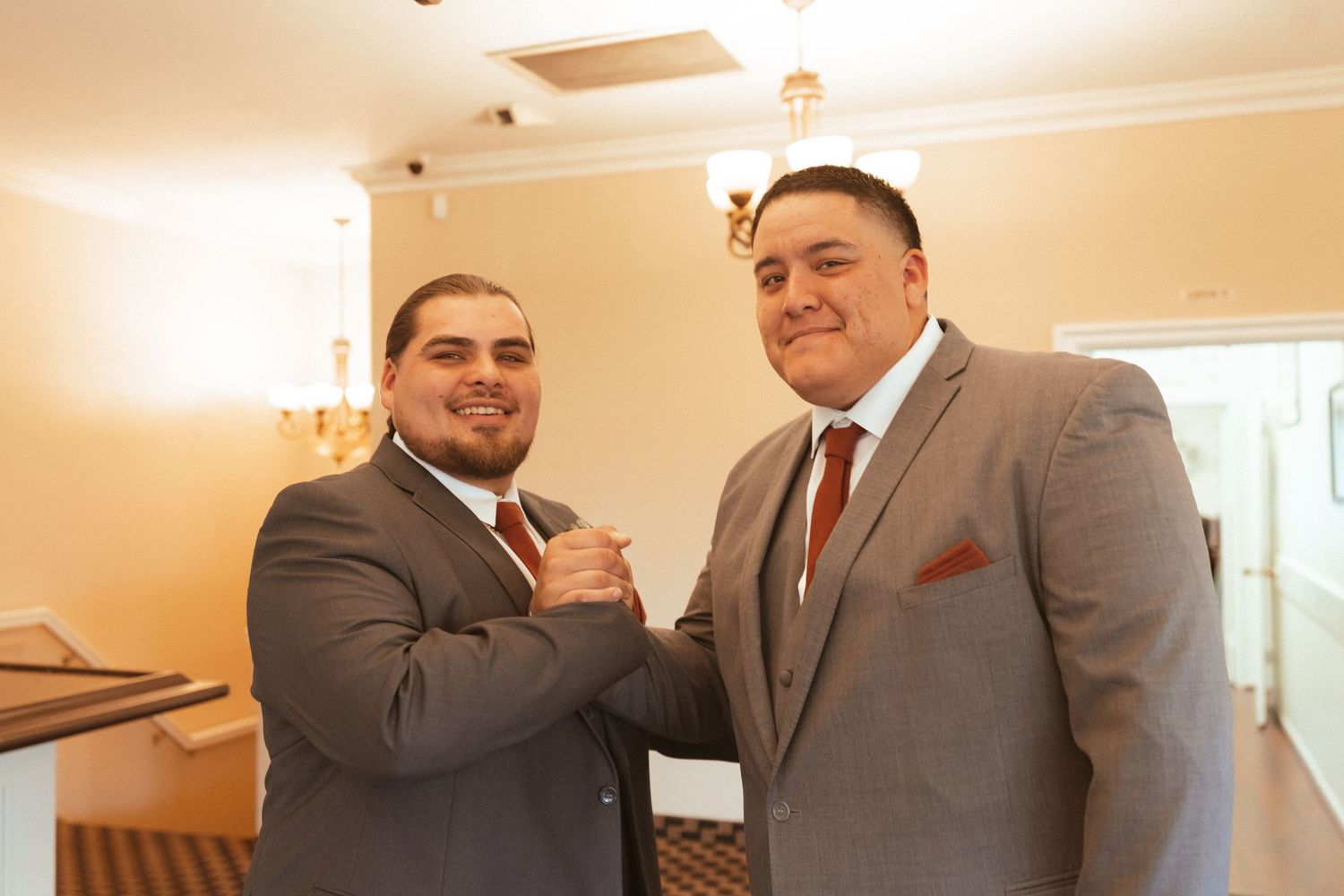Two men in formal attire share a celebratory moment while wearing matching gray suits and burgundy ties.