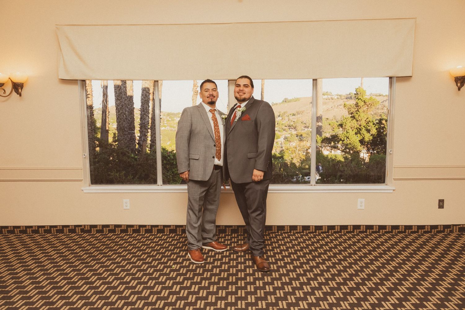 Two people in gray suits posing together on patterned carpet in front of a window.
