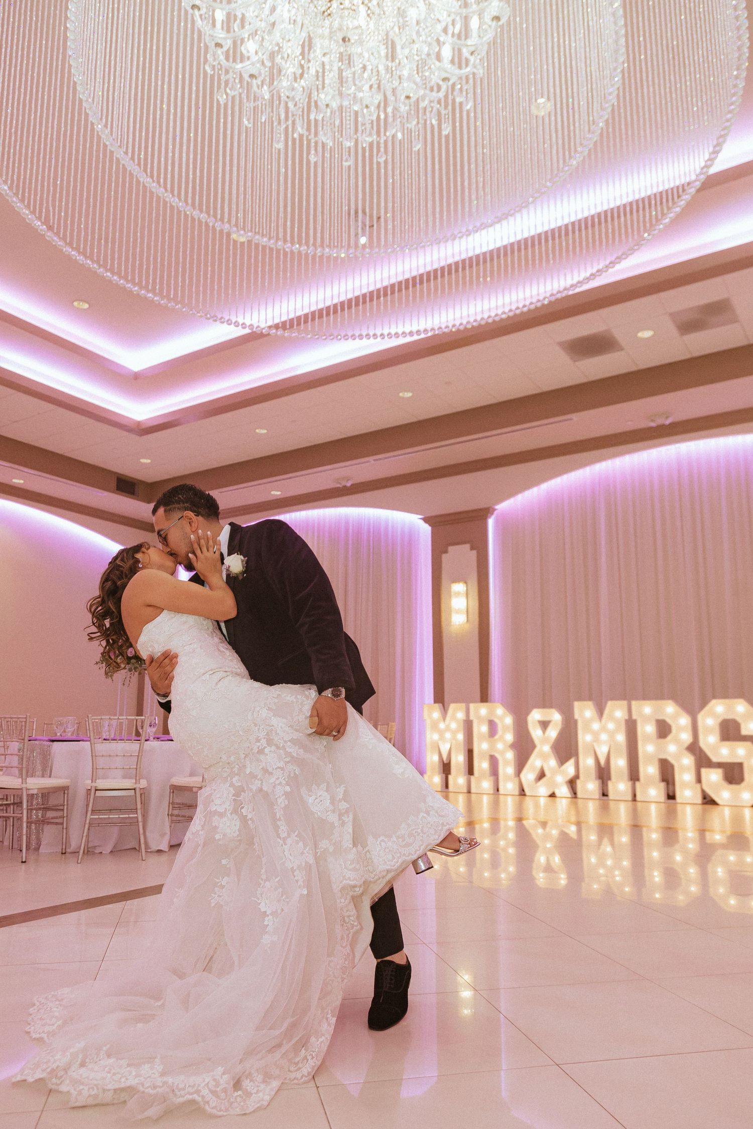 Wedding couple shares romantic dip kiss in pink-lit ballroom with illuminated MR & MRS sign.