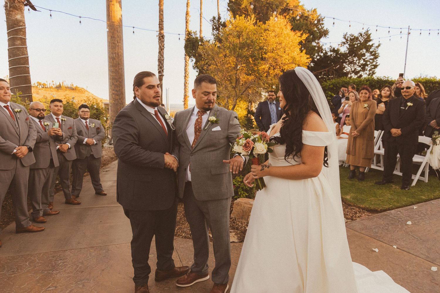Outdoor wedding ceremony with formal attire against backdrop of palm trees at sunset.
