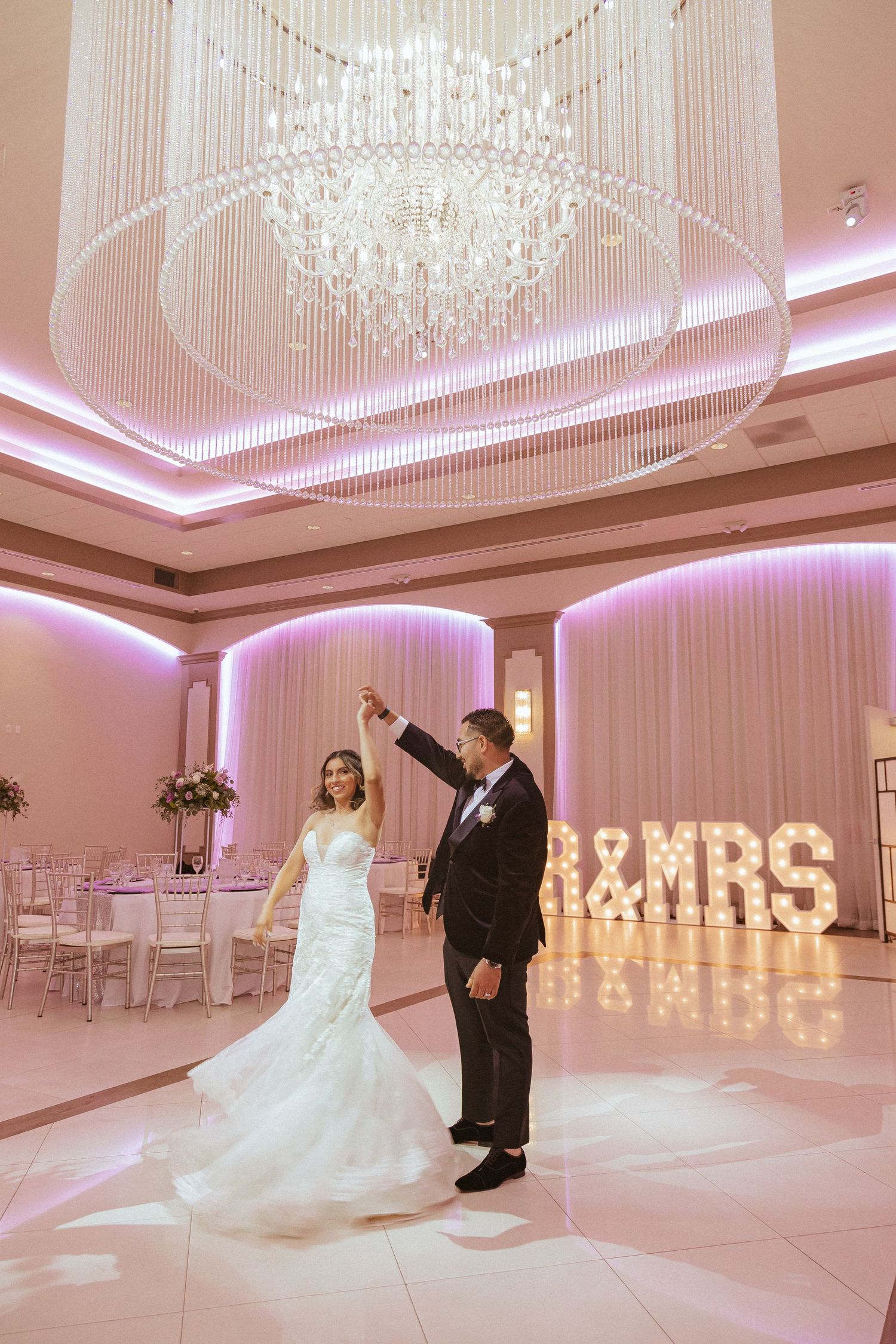 Elegant wedding dance captured under crystal chandelier with pink ambient lighting and MR&MRS marquee letters.
