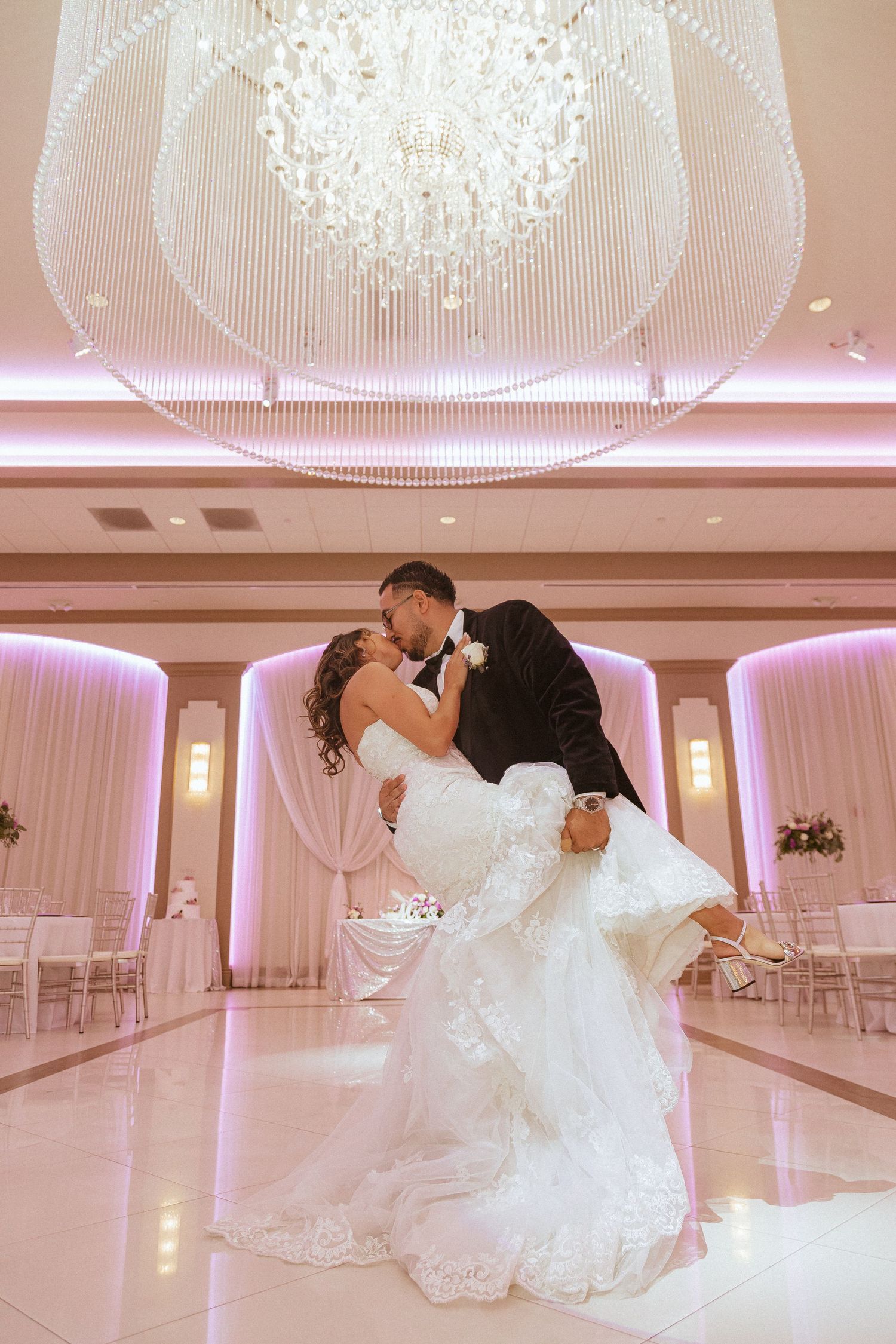 A romantic wedding couple shares a dip kiss under an elegant crystal chandelier in a pink-lit ballroom.