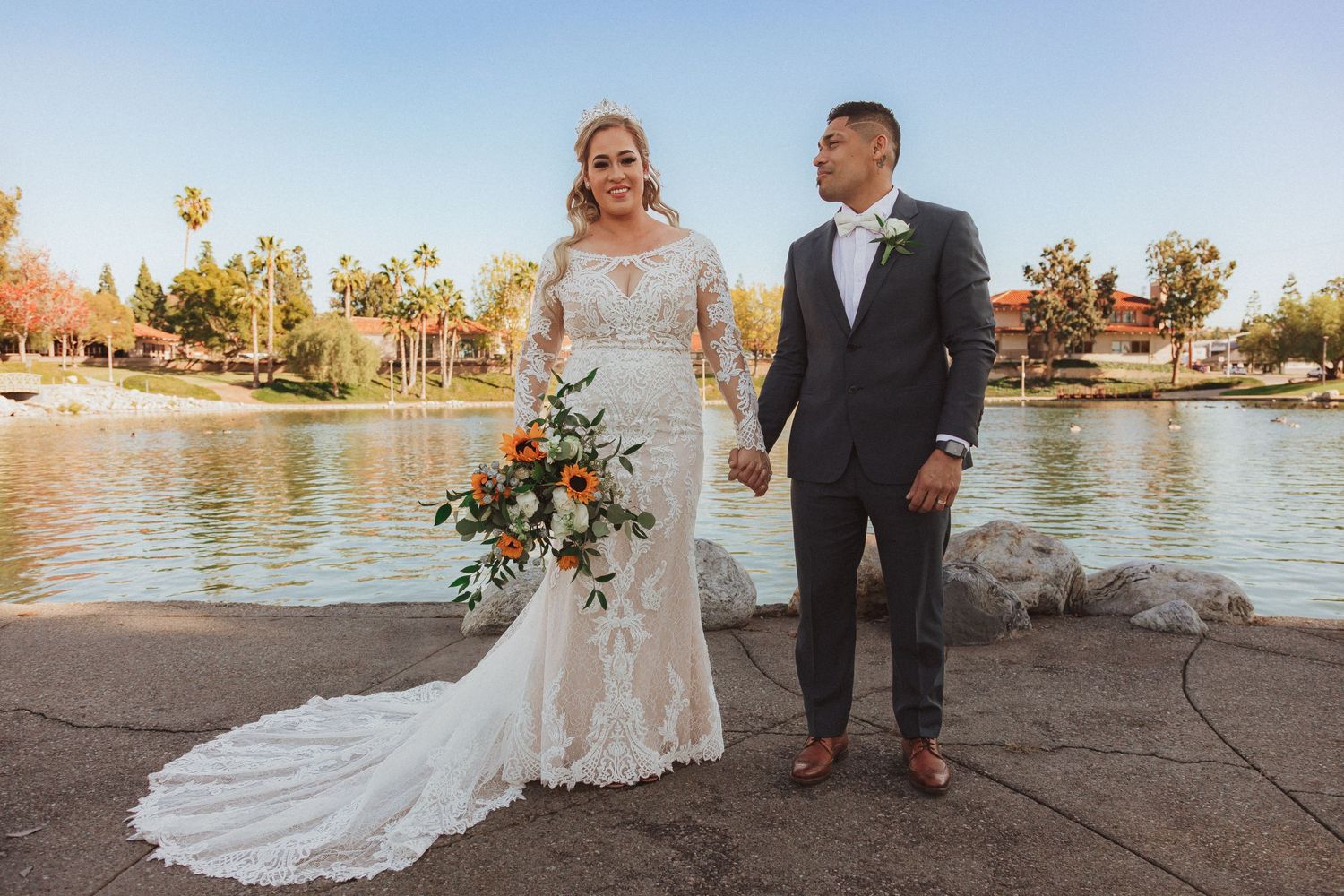 Bride with floral bouquet and groom pose together near scenic lake.