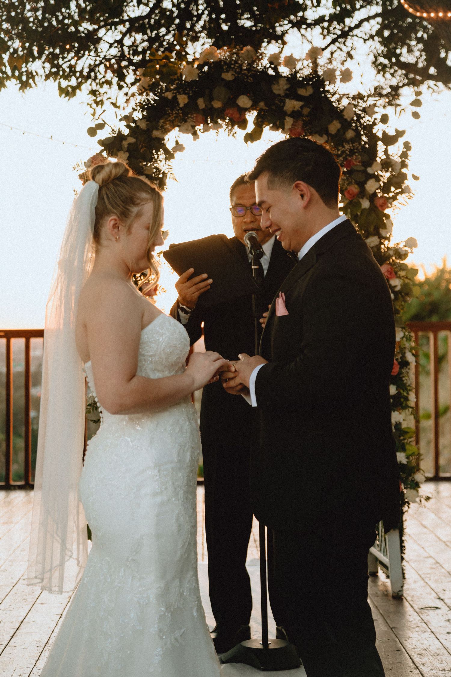 A wedding ceremony takes place beneath a floral arch with string lights at sunset.