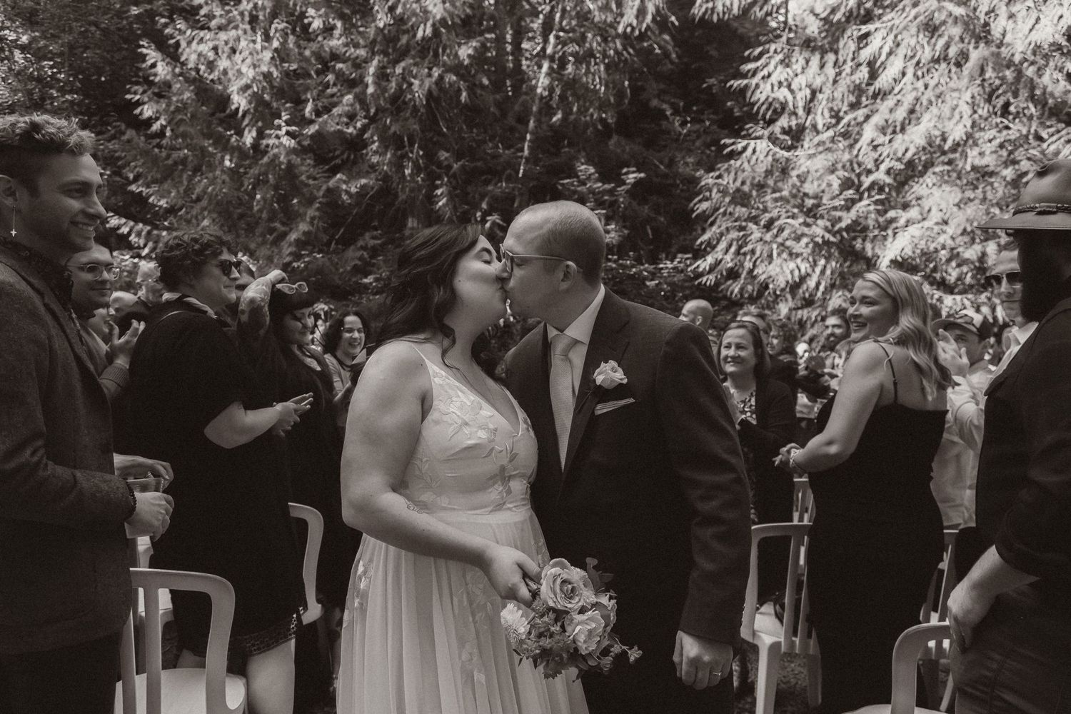 Black and white candid moment during forest wedding recessional with guests looking on.
