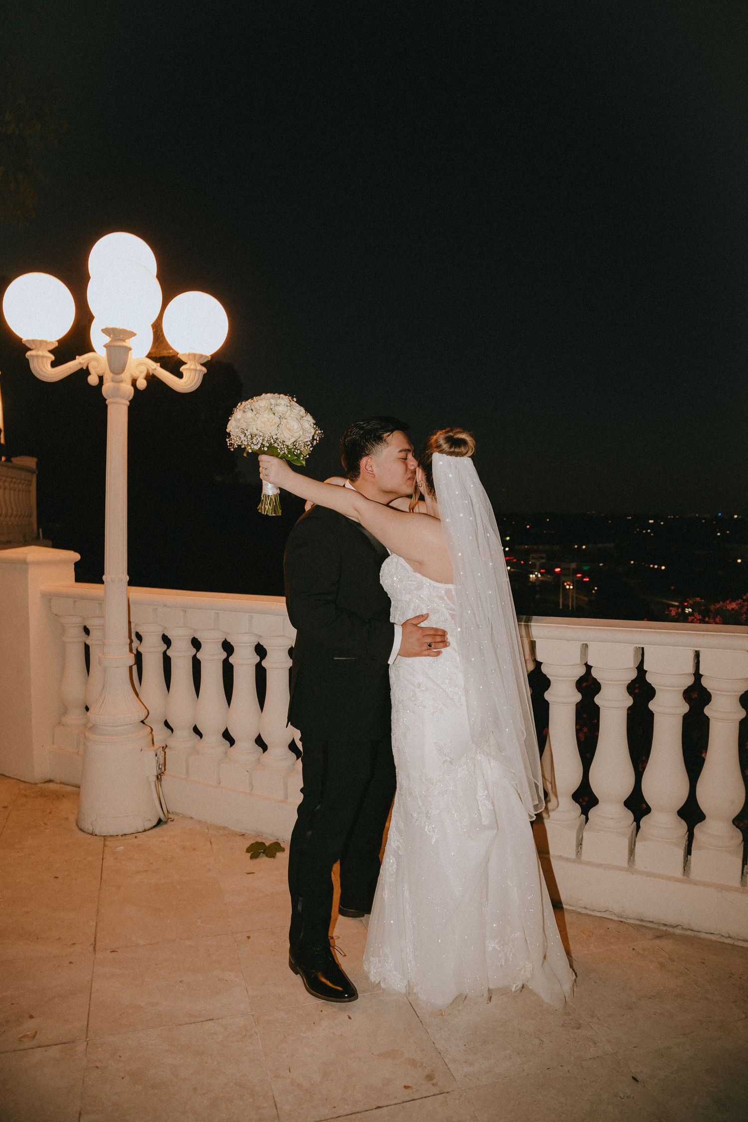 A tender moment between newlyweds dancing on a balcony with glowing vintage streetlamps.