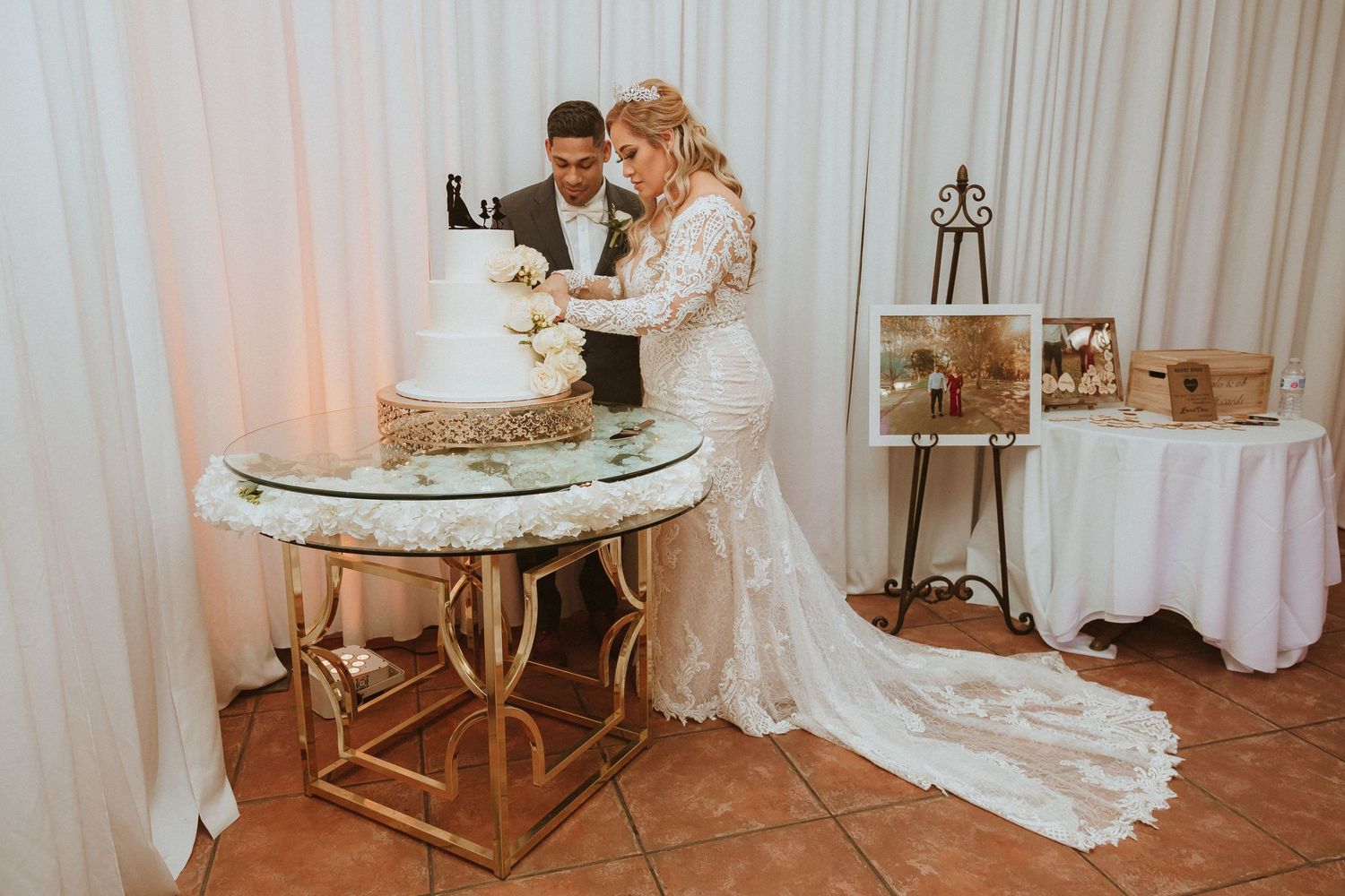 Bride in lace gown and groom cutting wedding cake at decorated reception table.