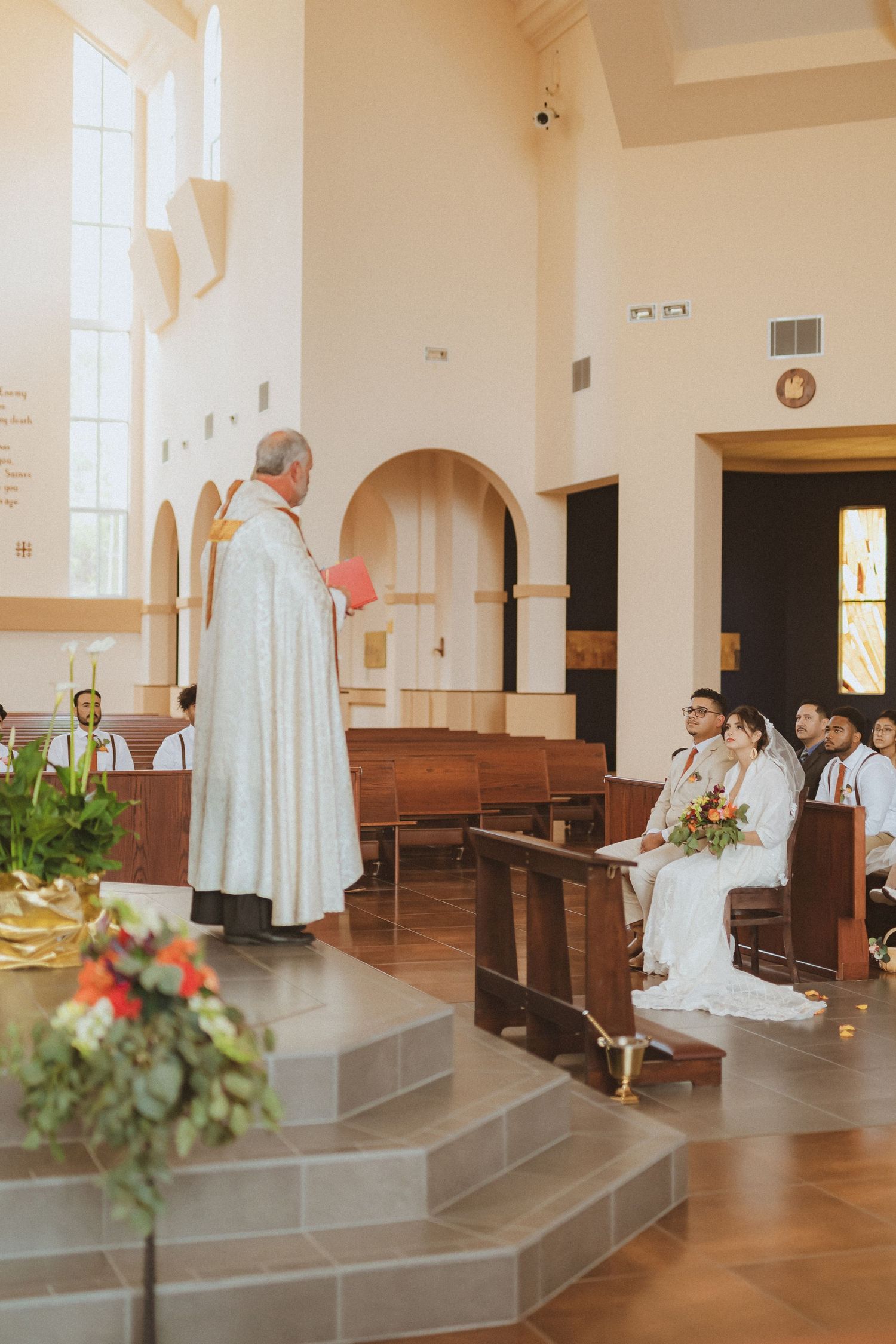 A Catholic priest in white robes conducts a wedding ceremony inside a church with arched windows and floral decorations.