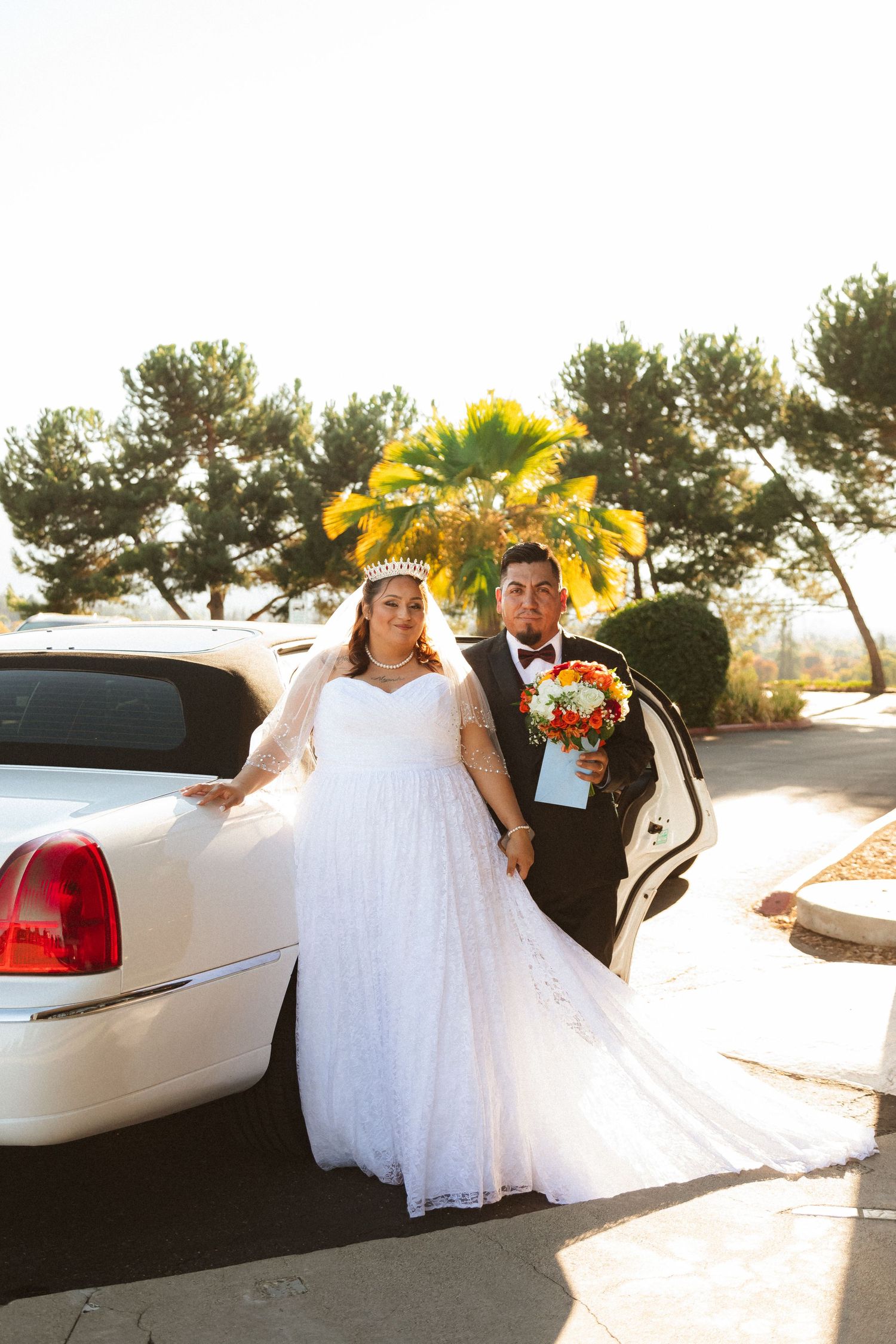 A couple poses by their white limousine after their wedding ceremony.