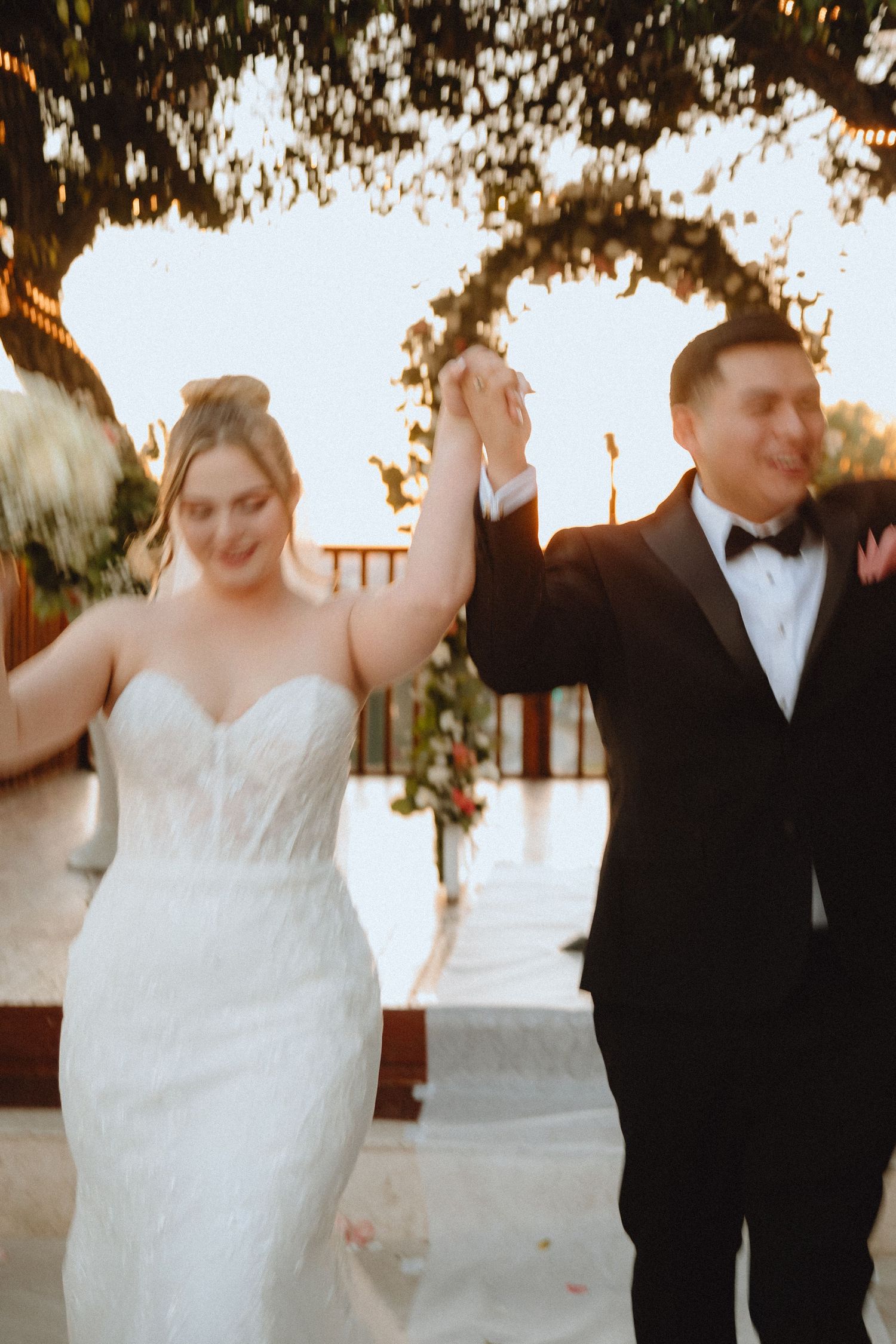 Wedding couple holds hands high in celebration under twinkling lights and trees.