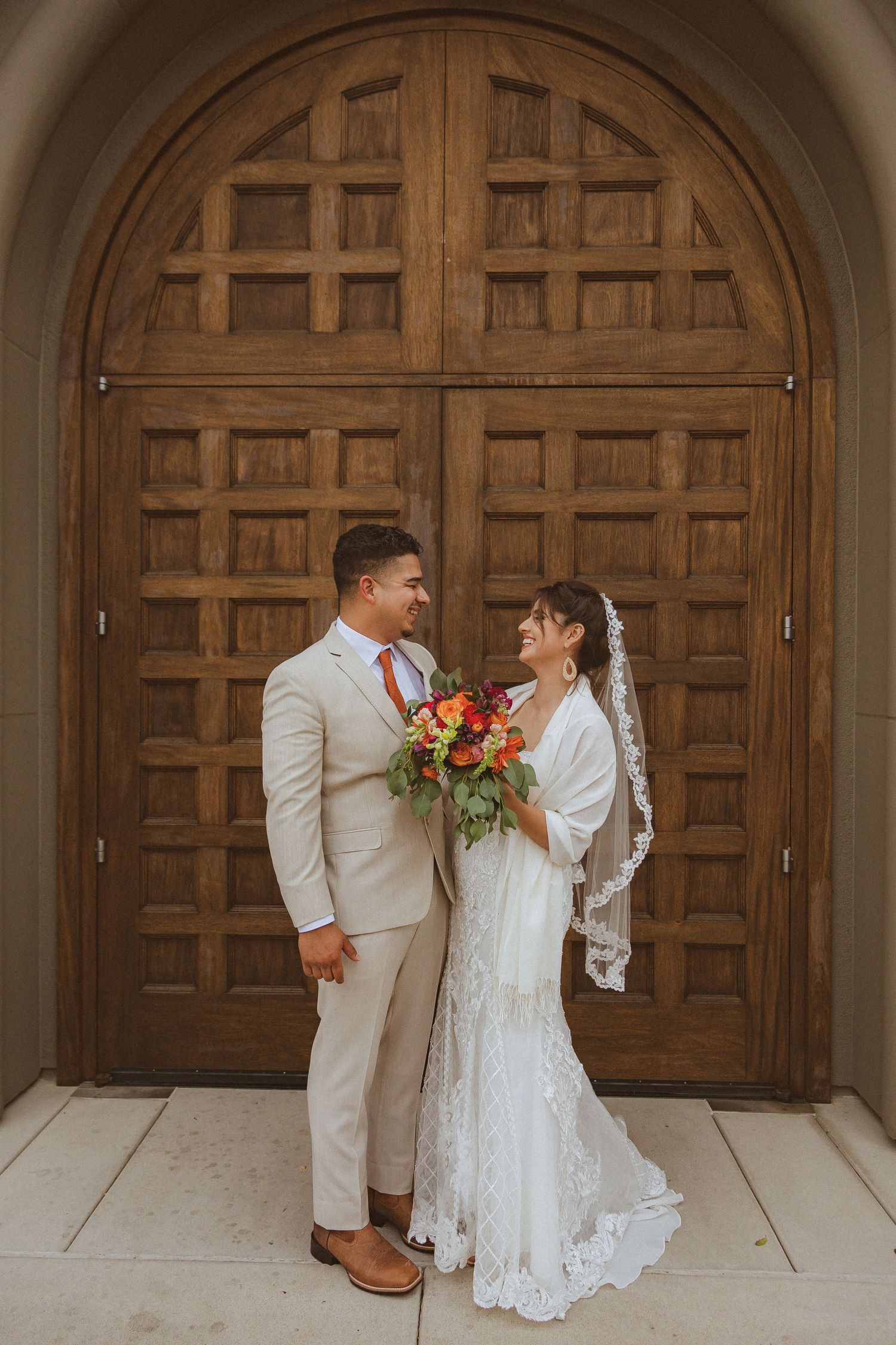Wedding portrait featuring light beige suit and lace dress against wooden church doors.